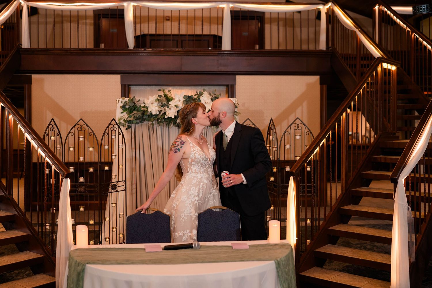 Newlyweds share a kiss at signing table between dual staircases in elegant indoor wedding venue.