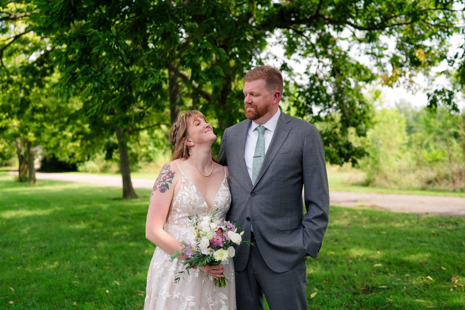 Bride in lace gown leans on groom in gray suit while holding bouquet in green park.