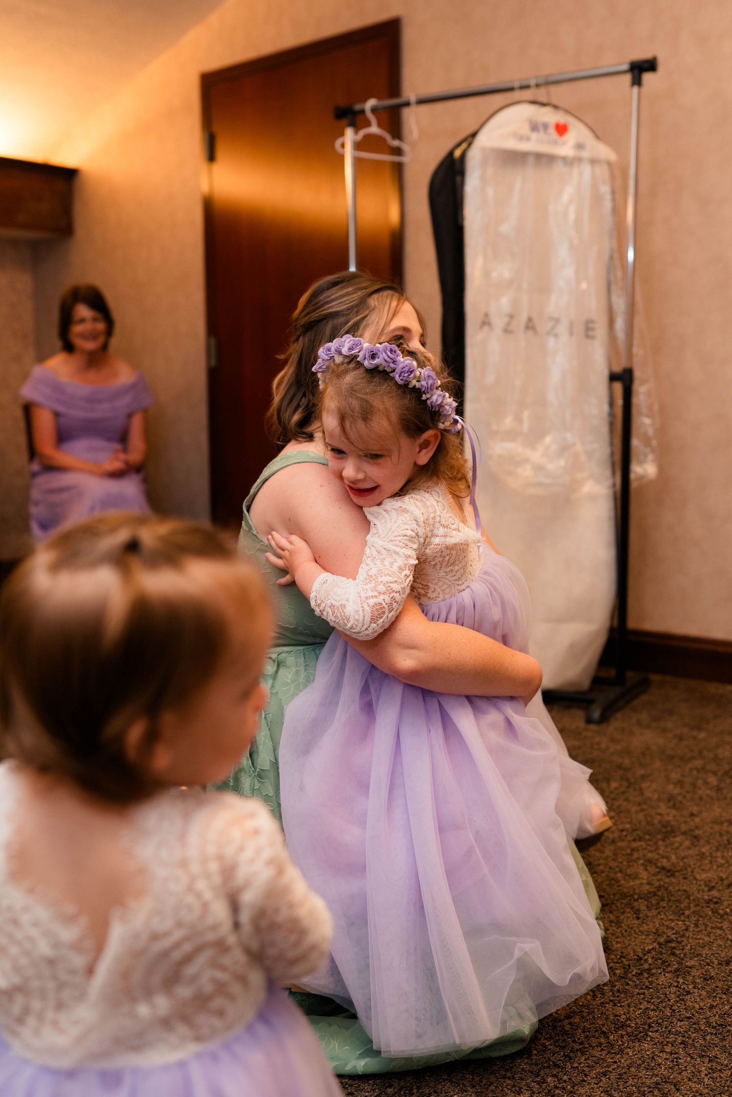 Young flower girl in purple tulle dress hugging bridesmaid while another flower girl watches in hotel room.