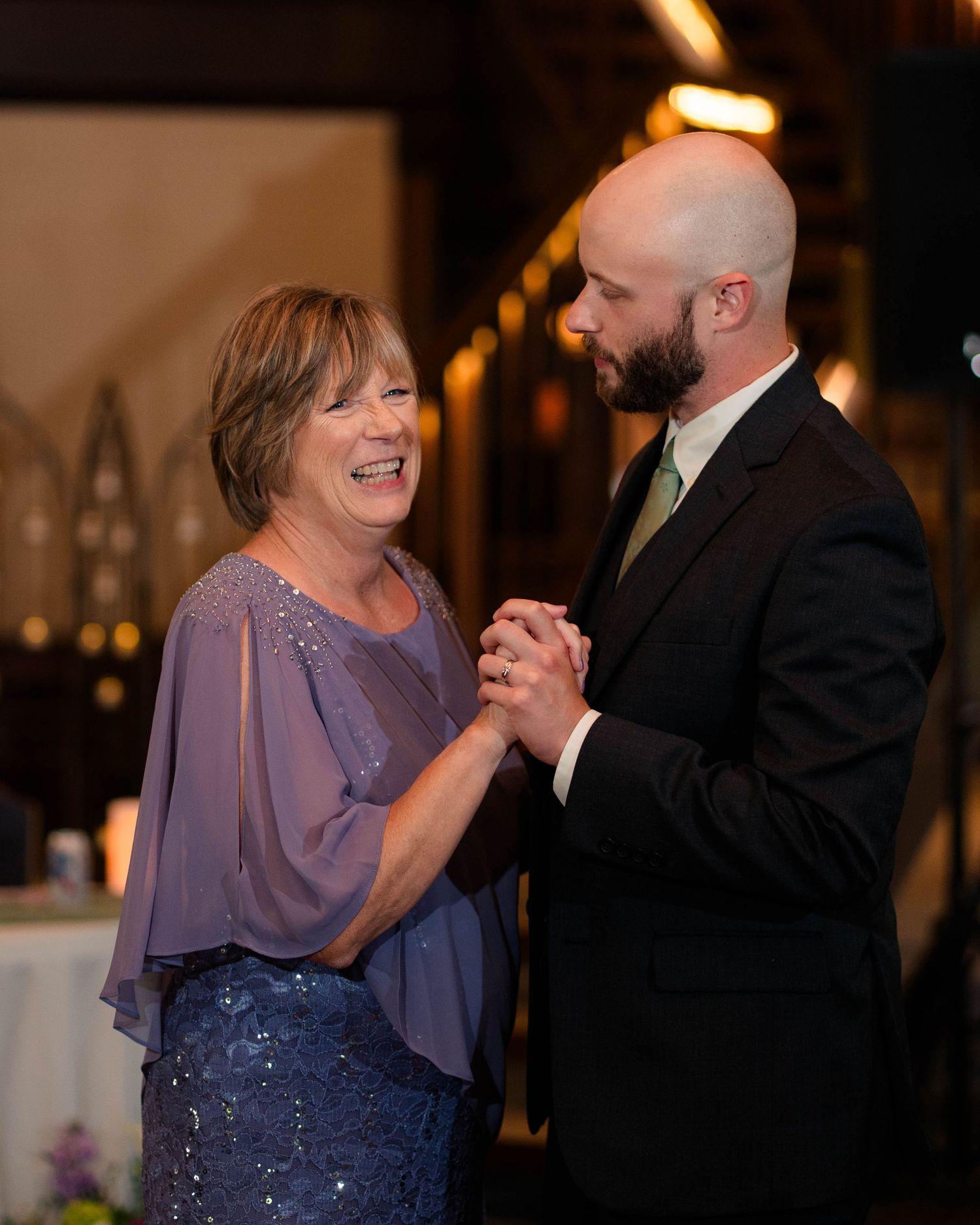 Groom shares mother-son dance with smiling woman in purple dress at wedding reception.