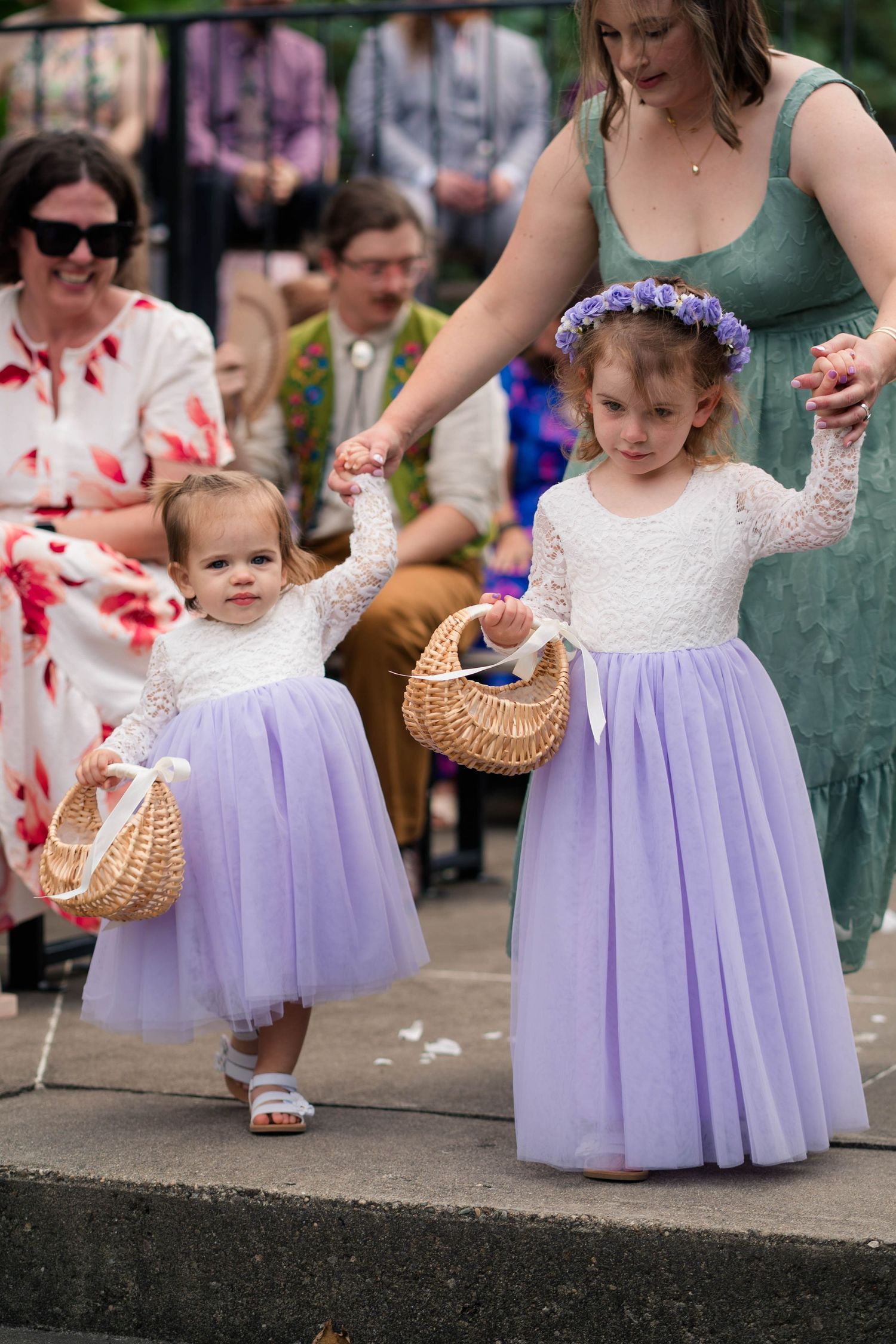 Two young flower girls in white lace and lavender tulle dresses carry baskets down the wedding aisle.