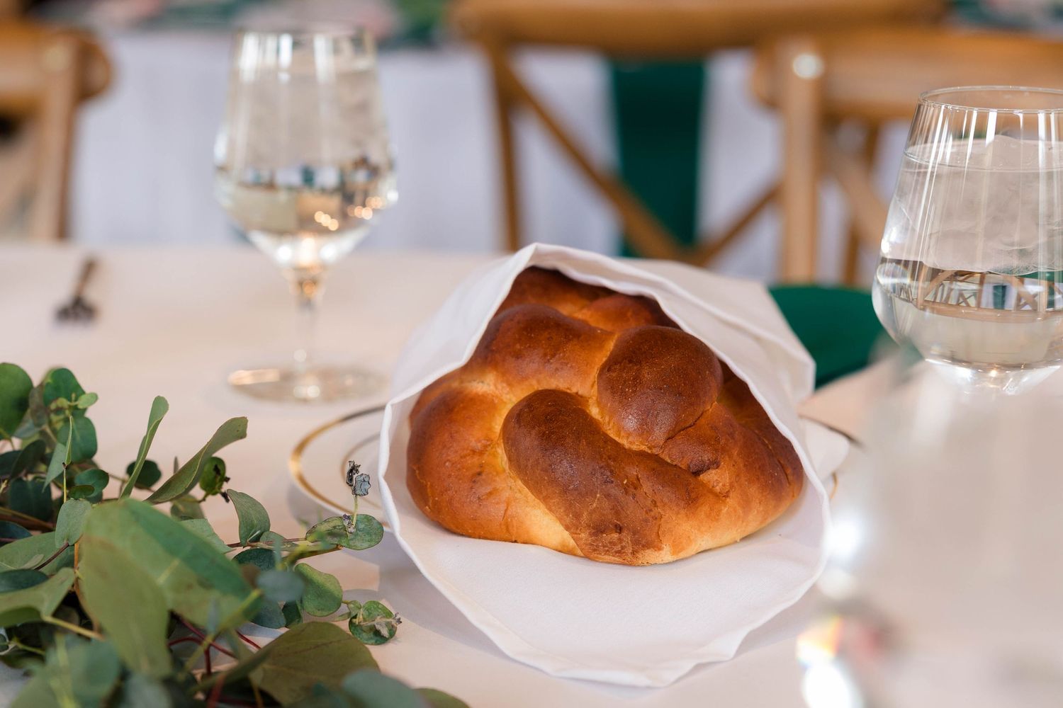 Freshly baked challah bread wrapped in white linen displayed on a gold-rimmed plate at a wedding table.