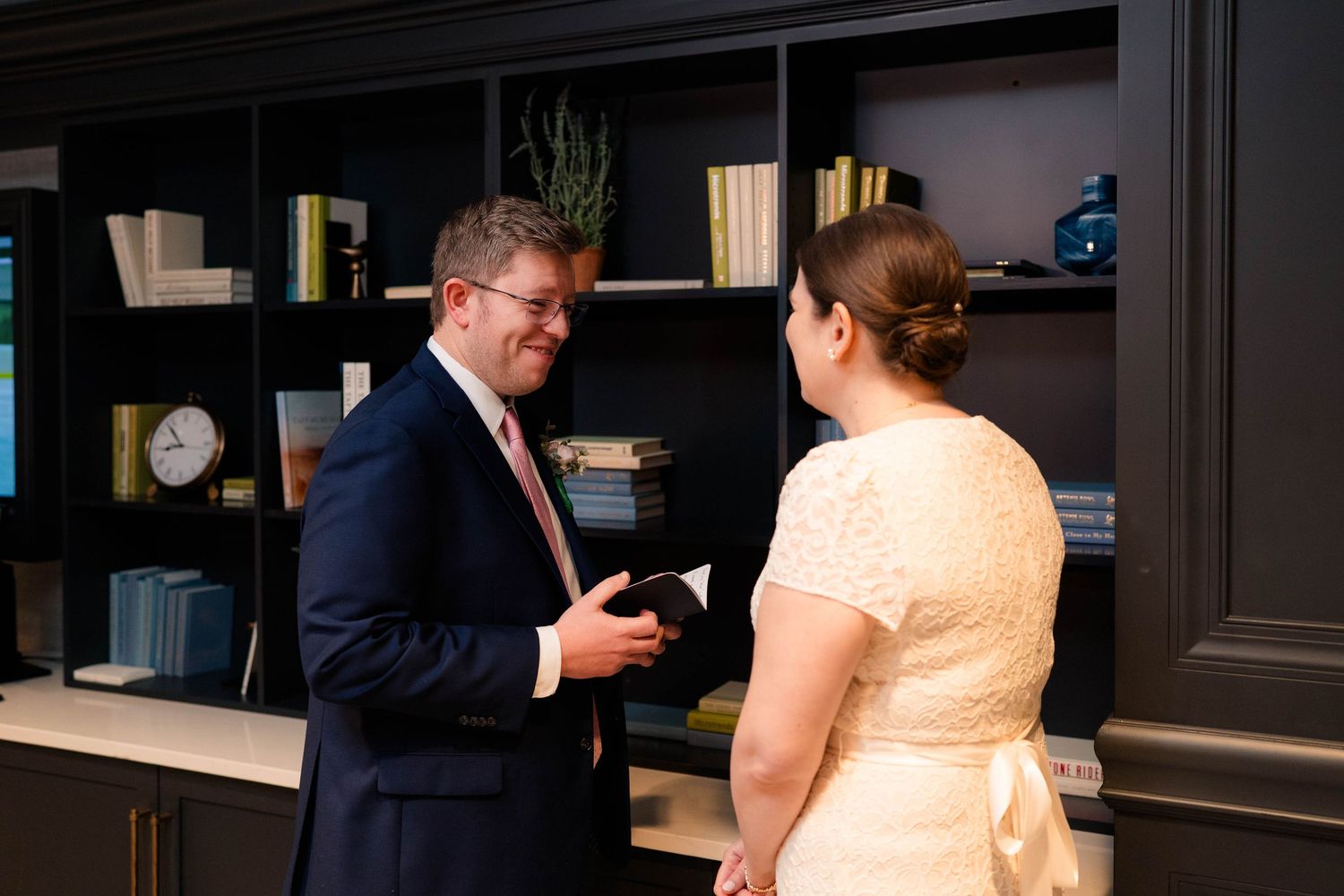 Bride and groom exchange private vows while holding cards in front of dark built-in bookshelves.