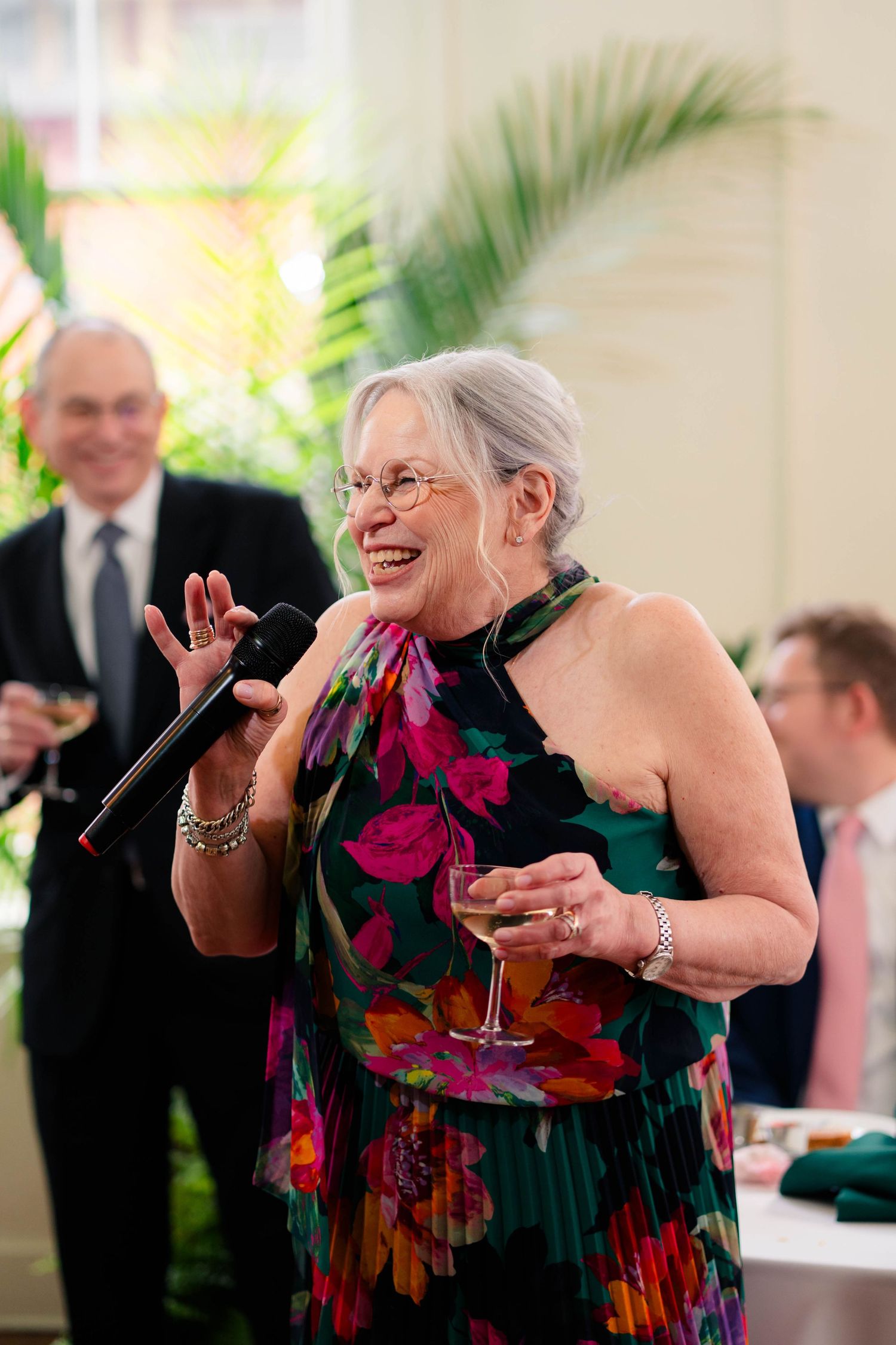 Older woman in colorful floral dress laughs while giving toast with microphone and wine.