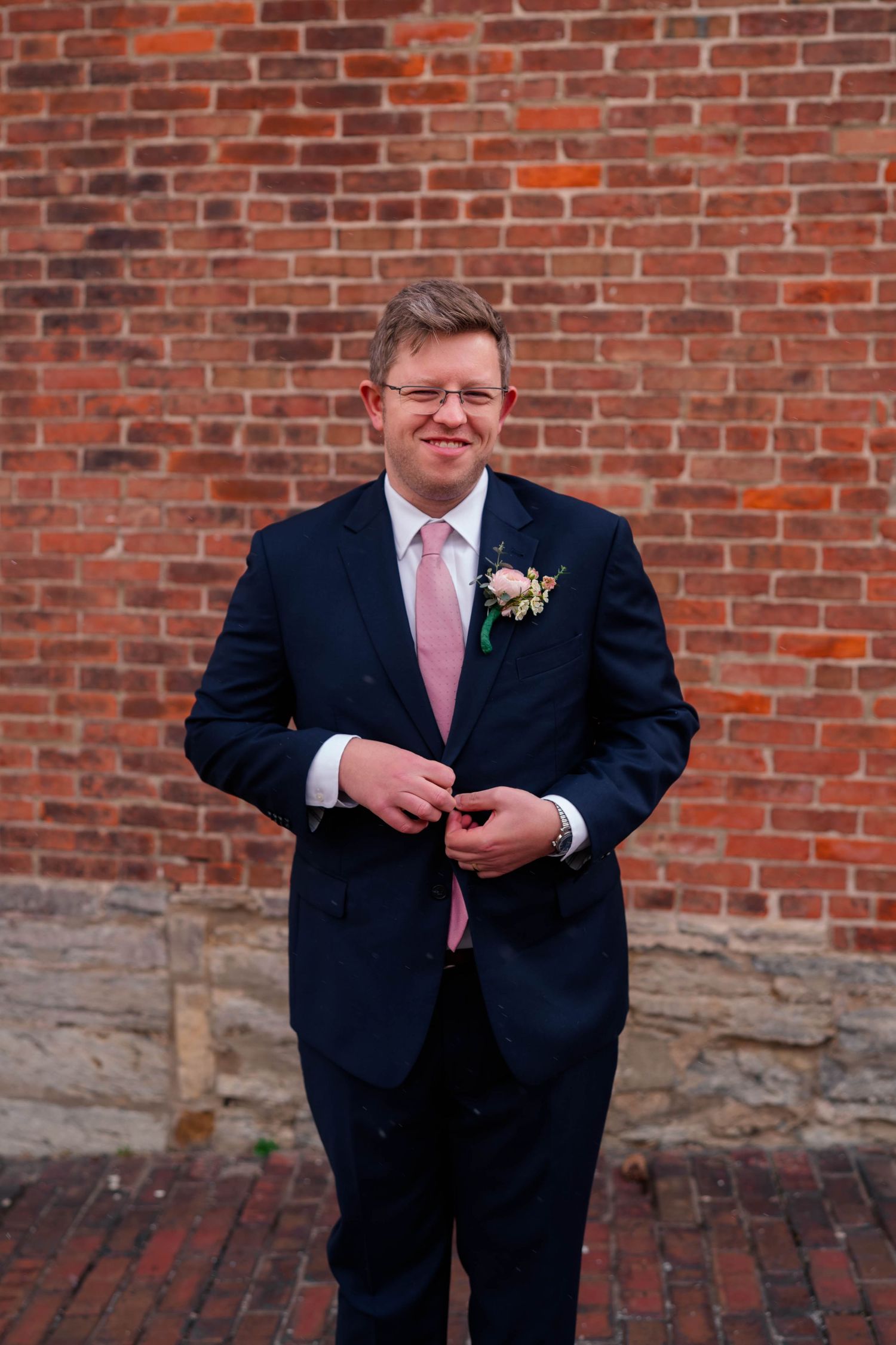Groom in navy suit with pink tie and boutonniere smiling in front of a red brick wall.