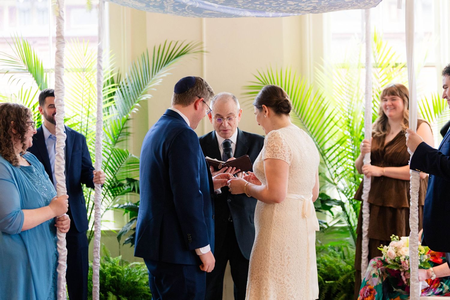 Couple exchanges rings under chuppah with palm trees and officiant during Jewish wedding.