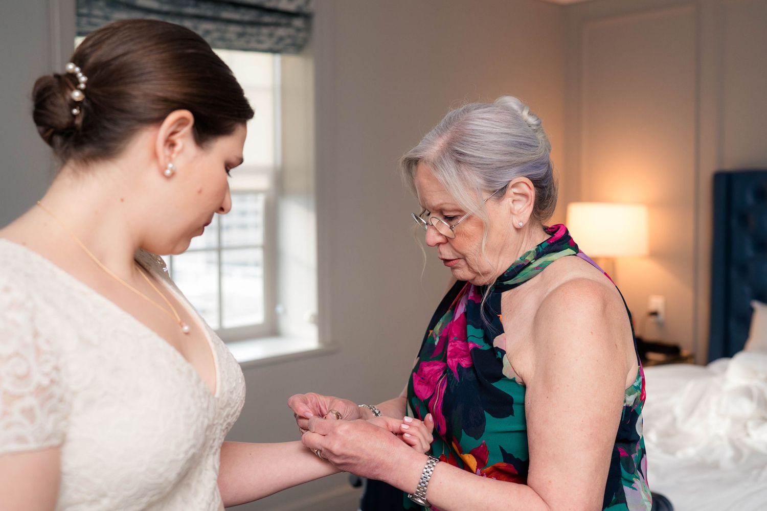 Mother helps bride fasten jewelry while standing in bright hotel room before wedding.