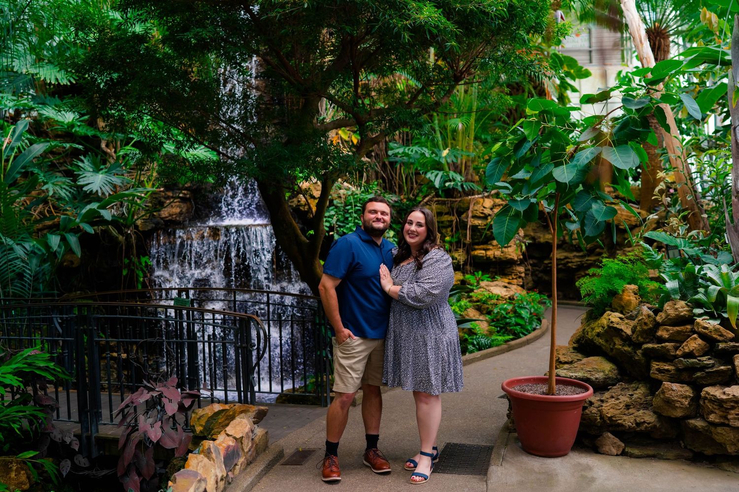 Couple smiling and holding hands in a lush indoor botanical garden with a waterfall backdrop.