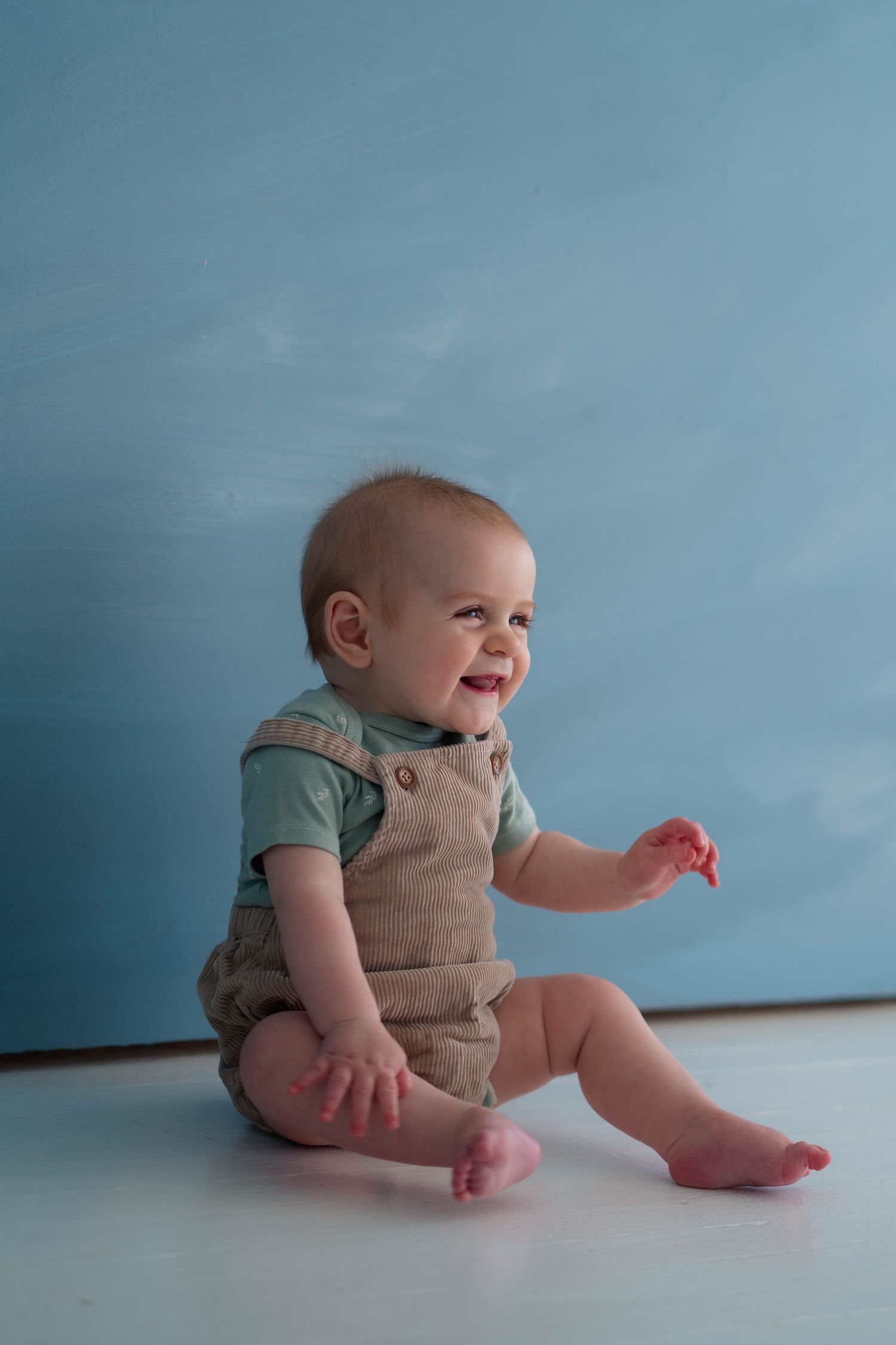 Laughing baby boy in tan overalls sitting on white floor against blue painted background.