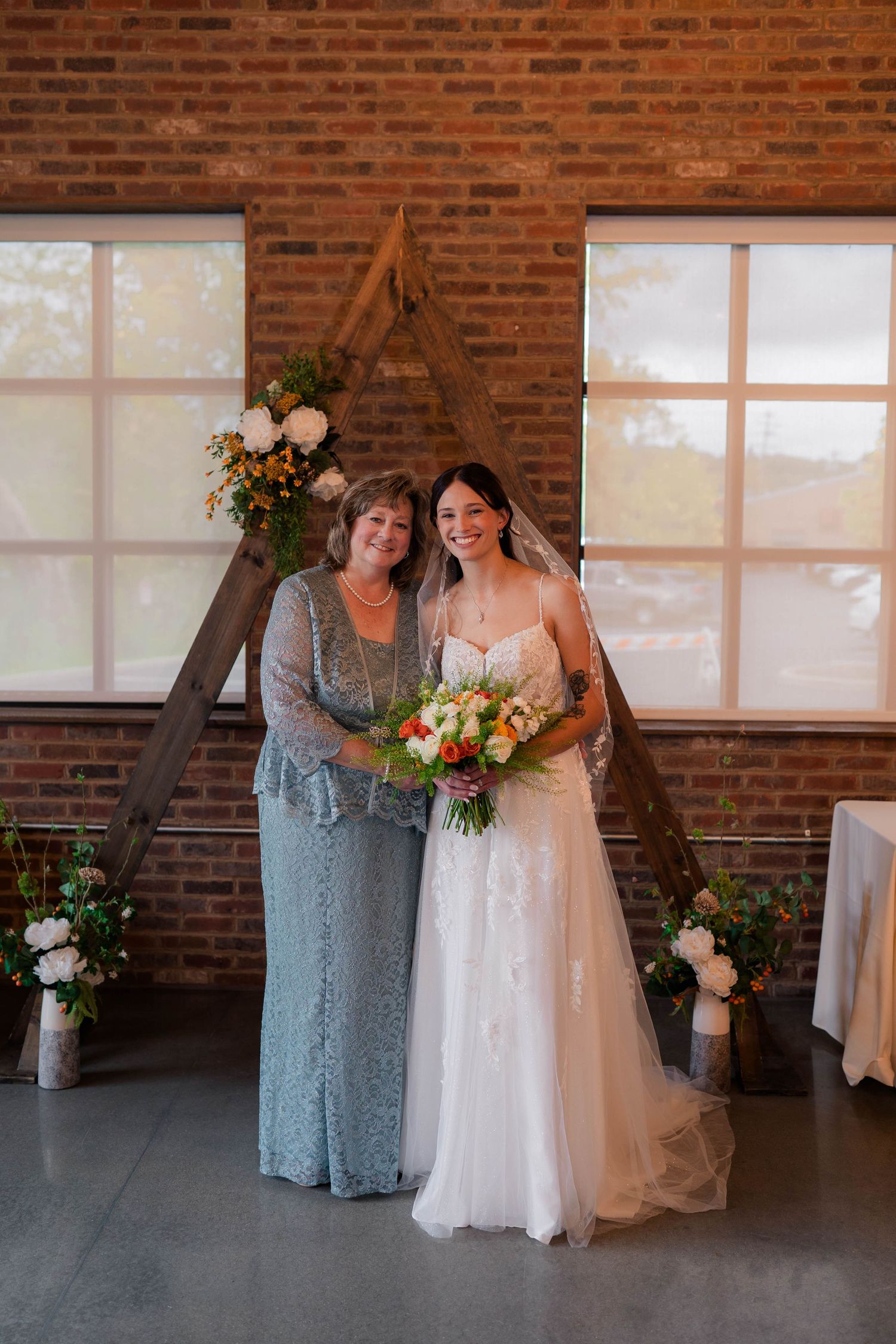 Bride in lace gown holding bouquet posing with older woman before wooden arch backdrop.