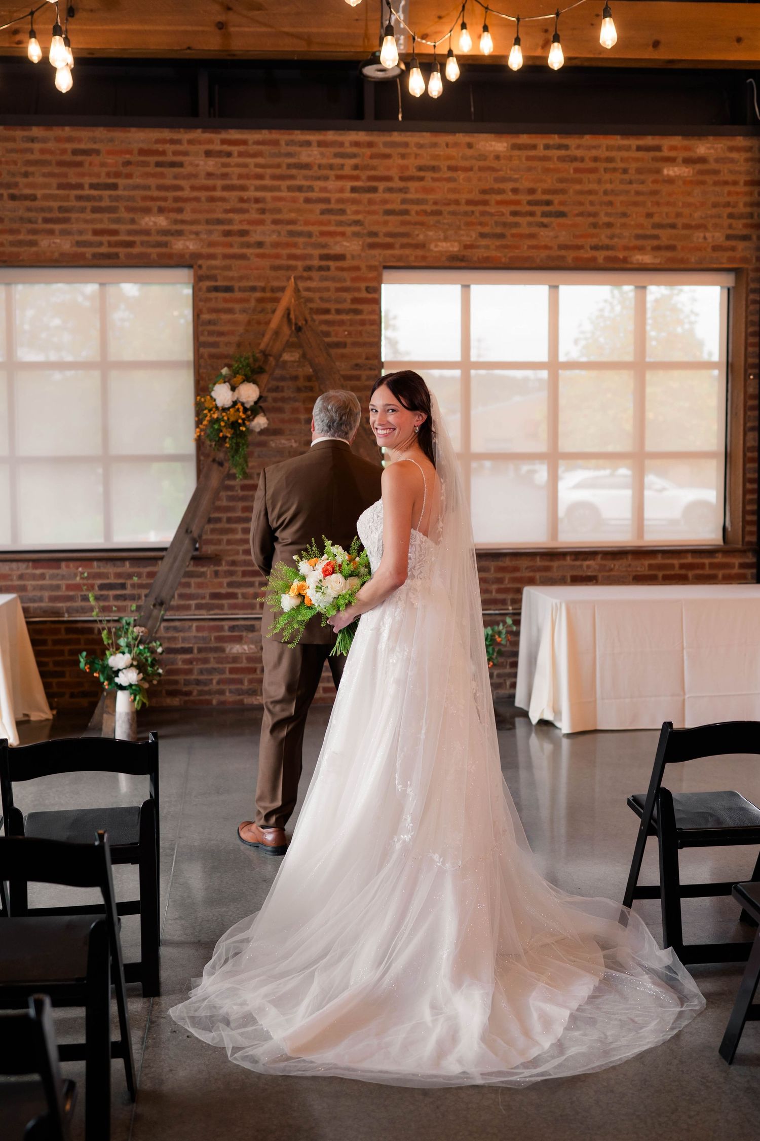 Smiling bride in white gown holding colorful bouquet stands in brick-walled wedding venue with Edison bulb lighting.