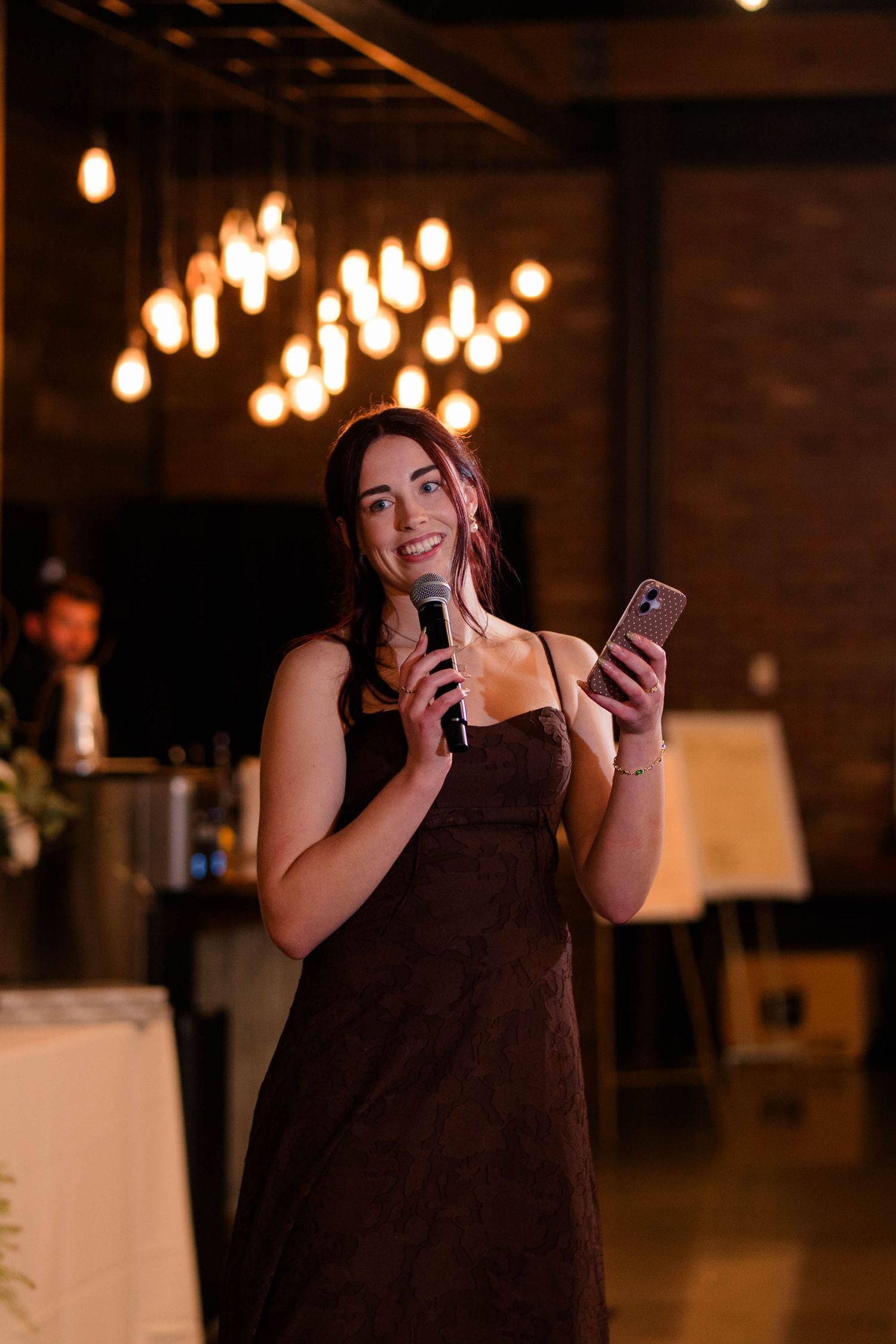 Bridesmaid in brown dress giving speech with microphone at wedding reception.