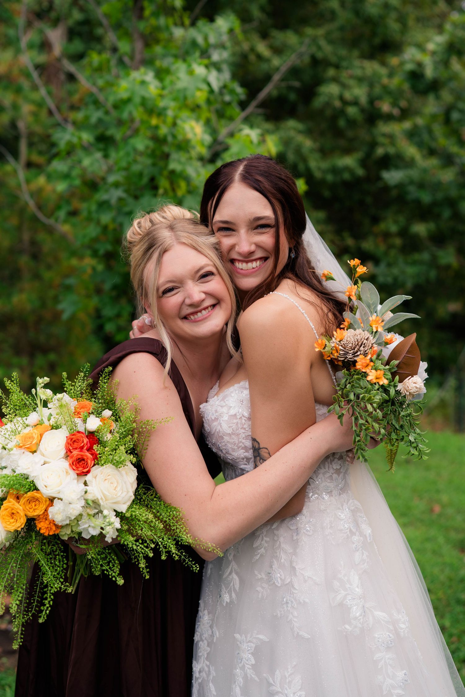 Bride in lace gown and bridesmaid in burgundy dress embrace smiling, both holding orange and white bouquets.