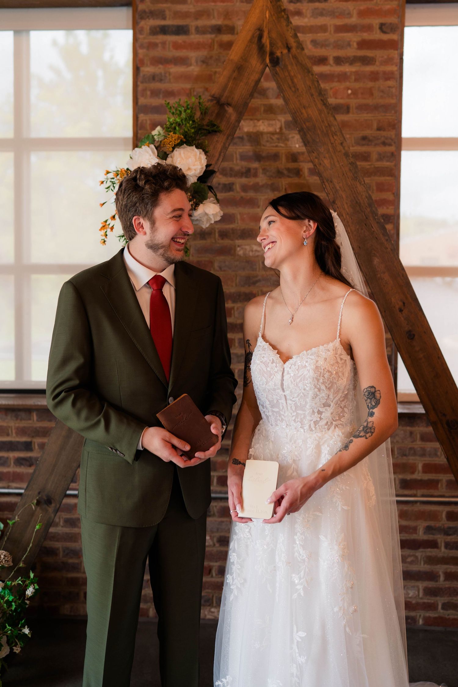 Bride and groom exchange vows holding books at rustic brick venue with wooden arch.
