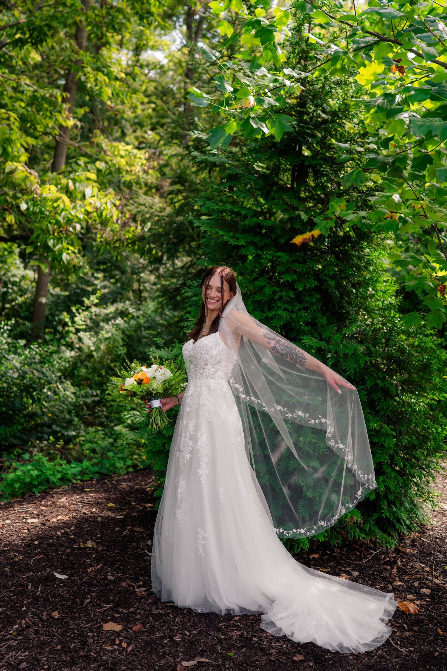 Radiant bride in lace gown twirling veil while holding bouquet in lush green garden.