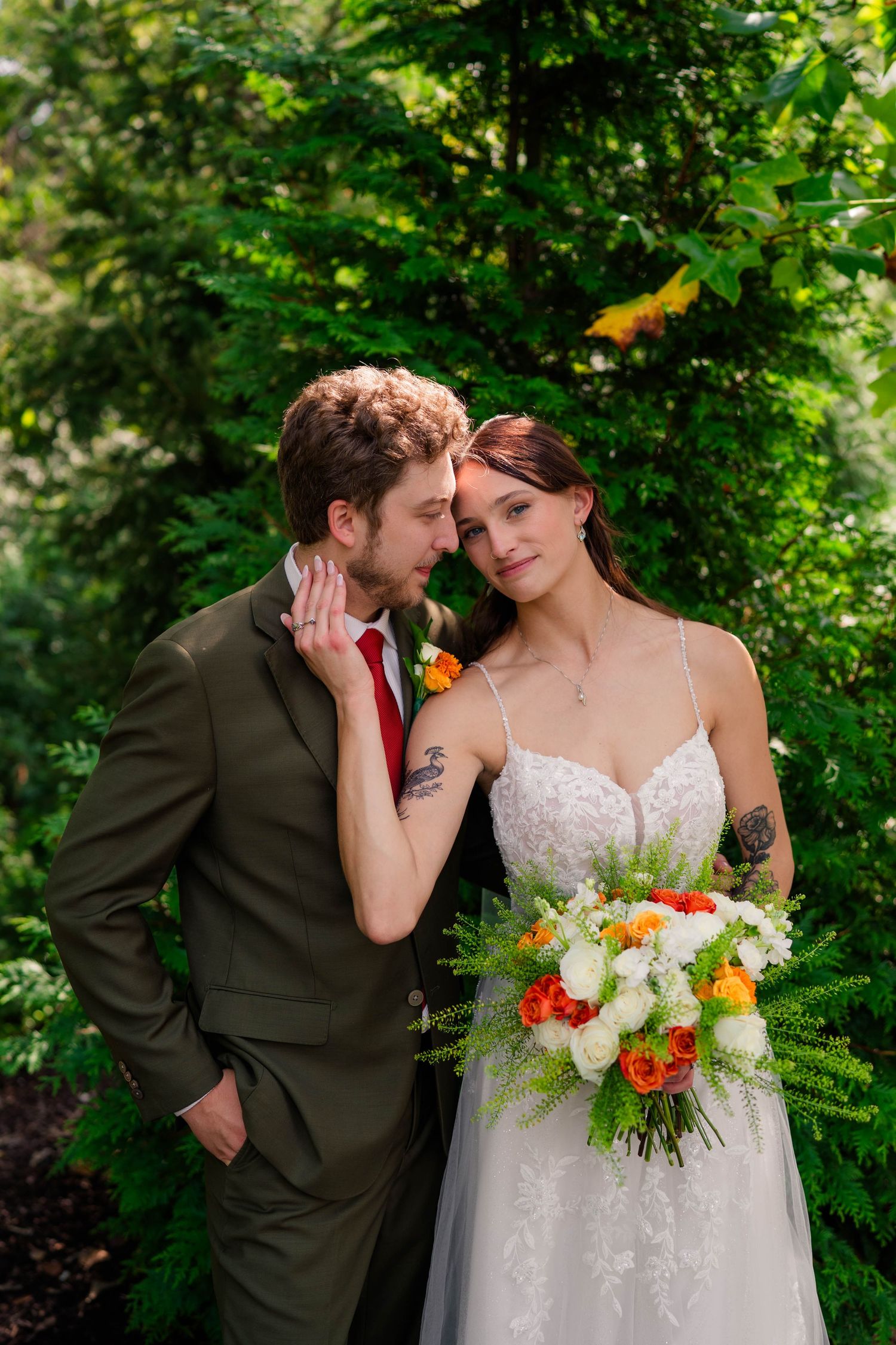 Bride and groom pose together outdoors among lush green trees, bride holding vibrant orange and white bouquet.