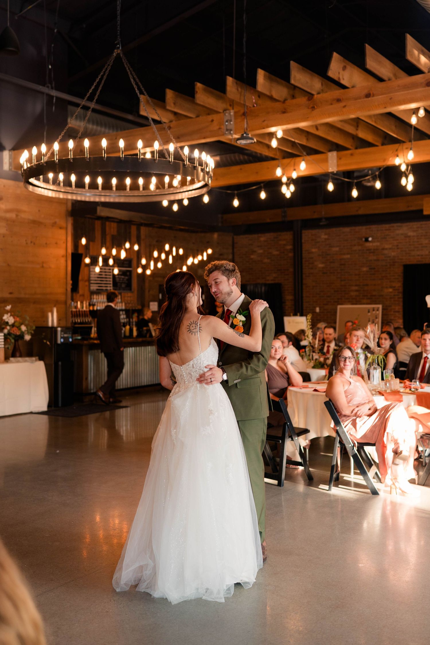 Newlyweds share first dance in warmly lit rustic reception hall with guests watching.