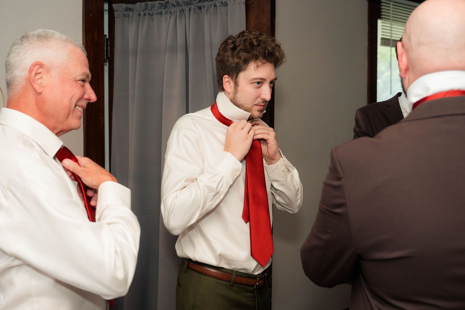 Three men in white shirts adjusting red ties, preparing for a formal event or wedding ceremony.