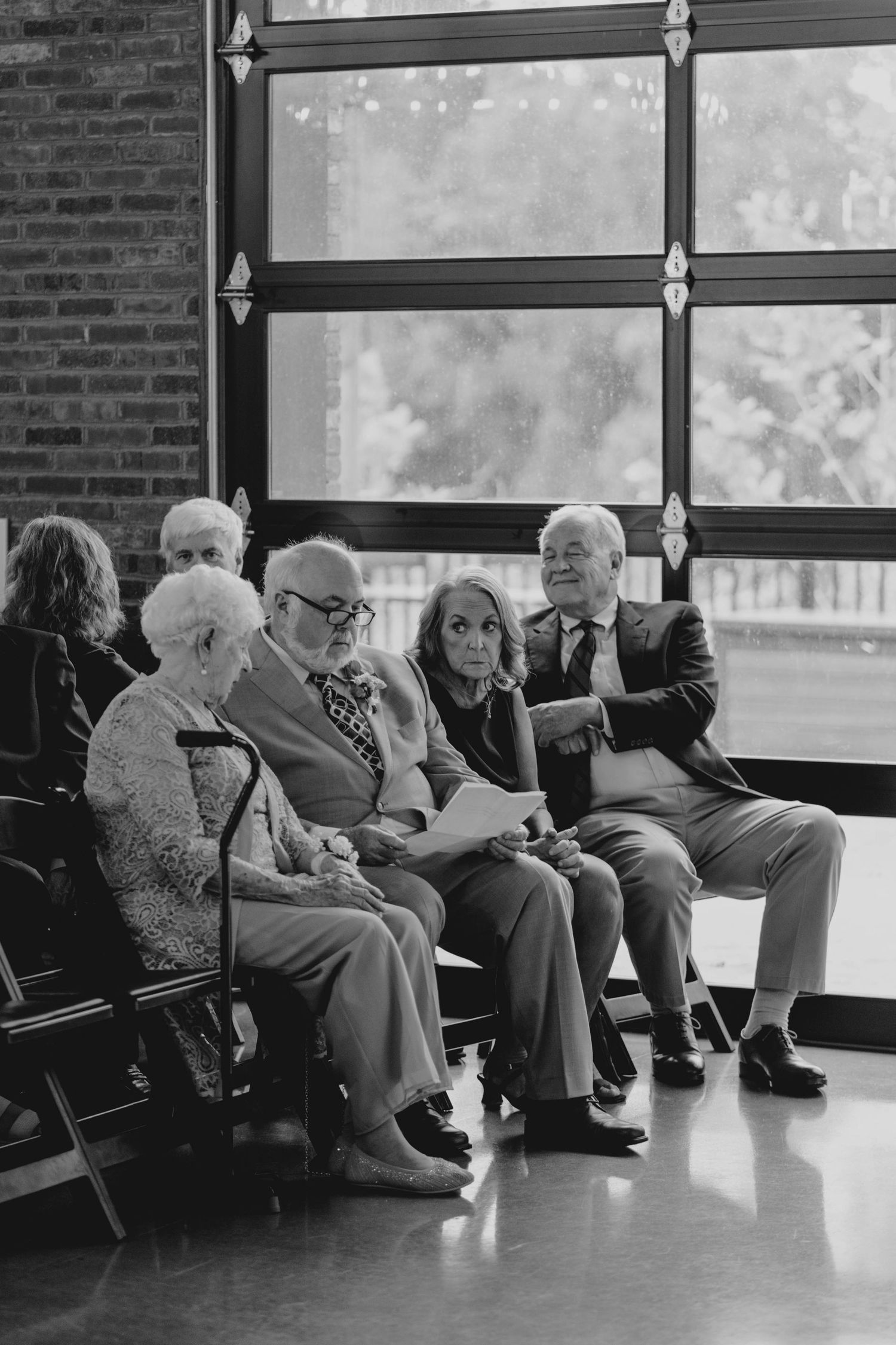 Black and white photo of elderly wedding guests seated and chatting near large windows at a ceremony.