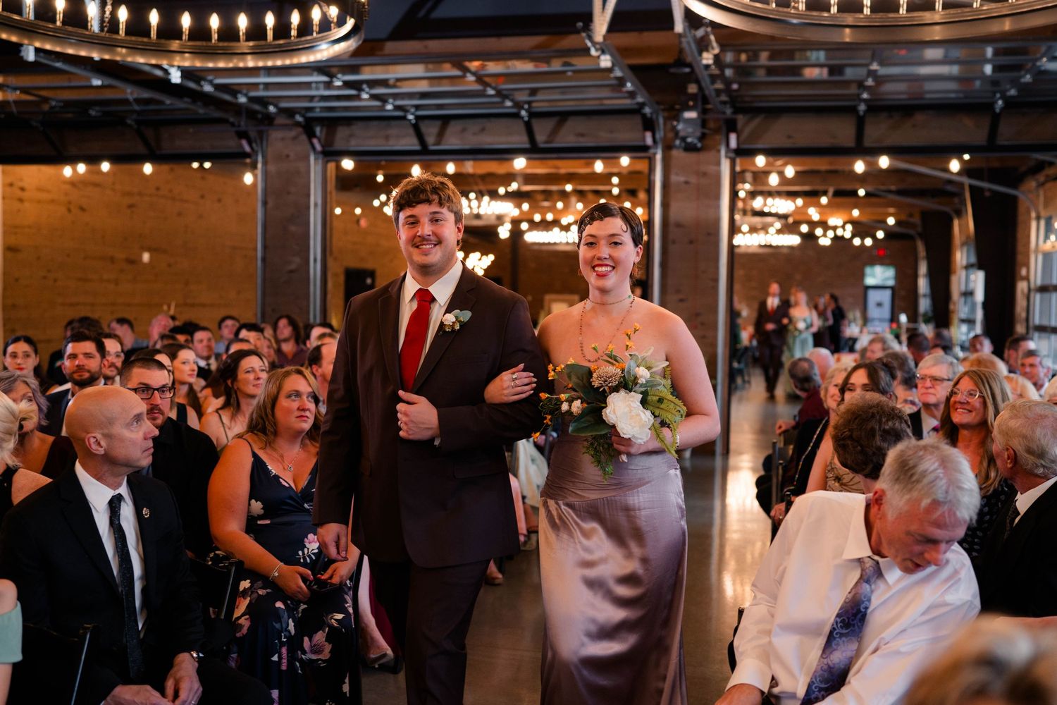Smiling bridesmaid in silver gown escorts groomsman down aisle at elegant indoor wedding ceremony.