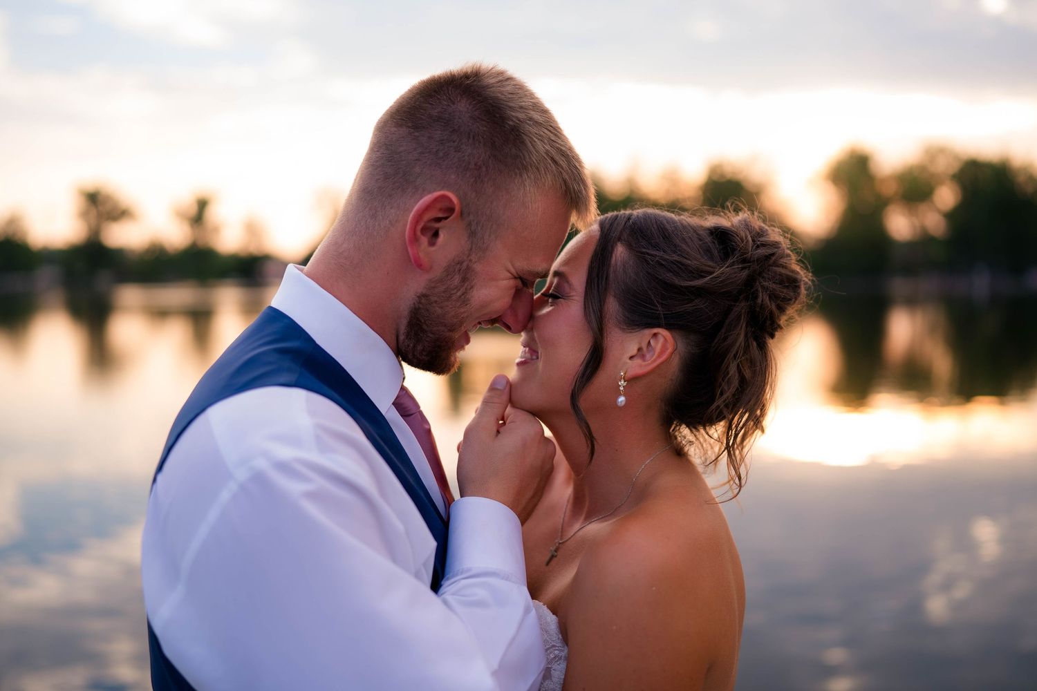Bride and groom share a romantic sunset moment by a reflective lake, touching foreheads tenderly on their wedding day.