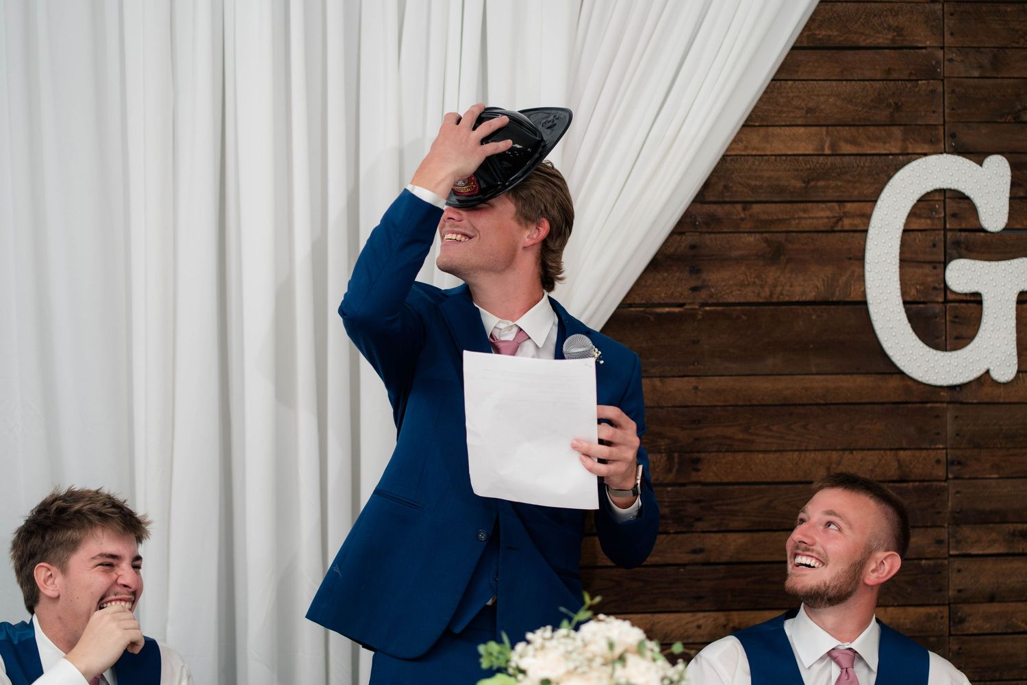 Groomsman delivers humorous wedding speech while holding paper and sunglasses at rustic reception venue.