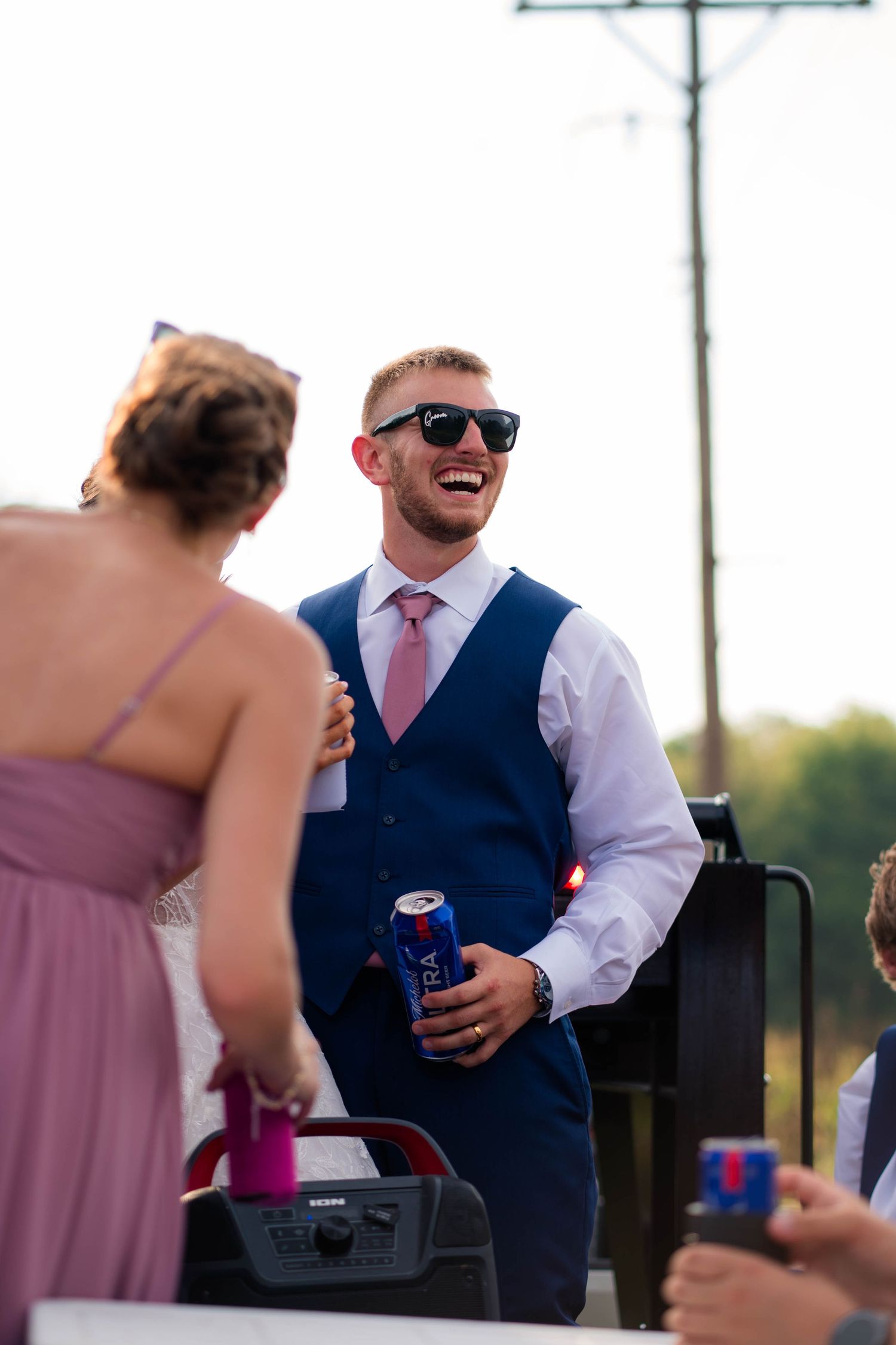 Laughing groom in navy vest and pink tie holds beverage can at outdoor wedding celebration.
