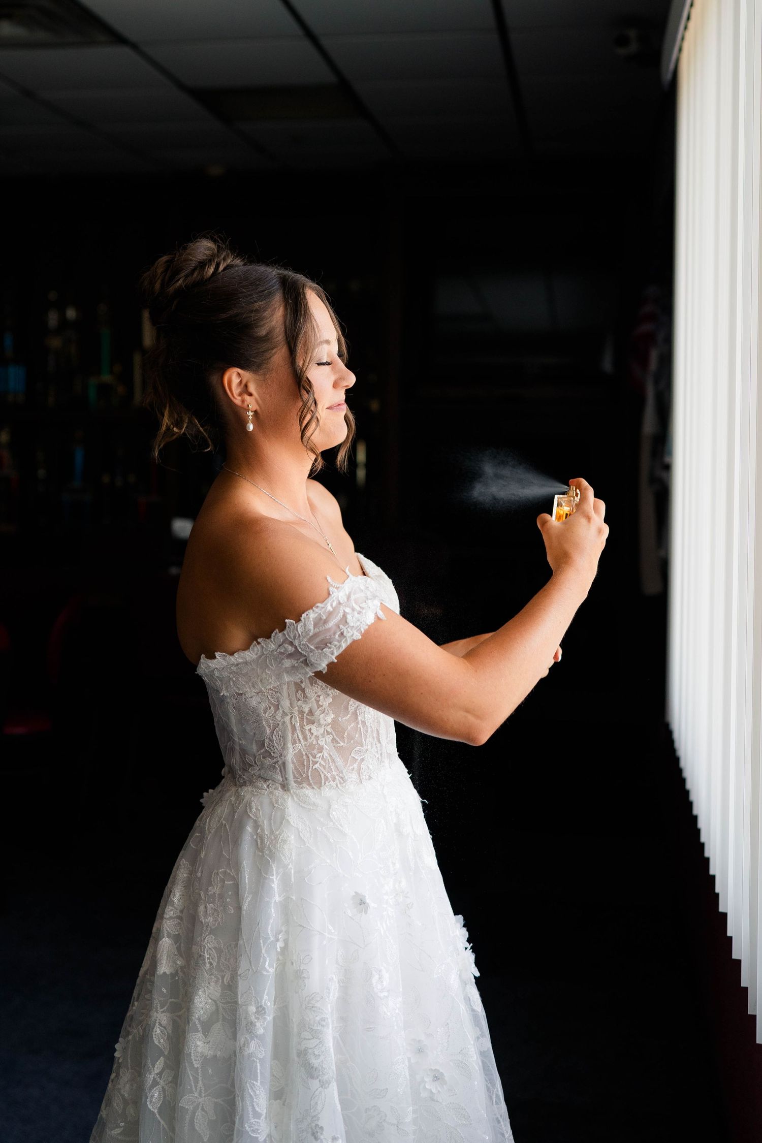 Bride in off-shoulder lace wedding dress applying perfume in dramatic side lighting before ceremony.