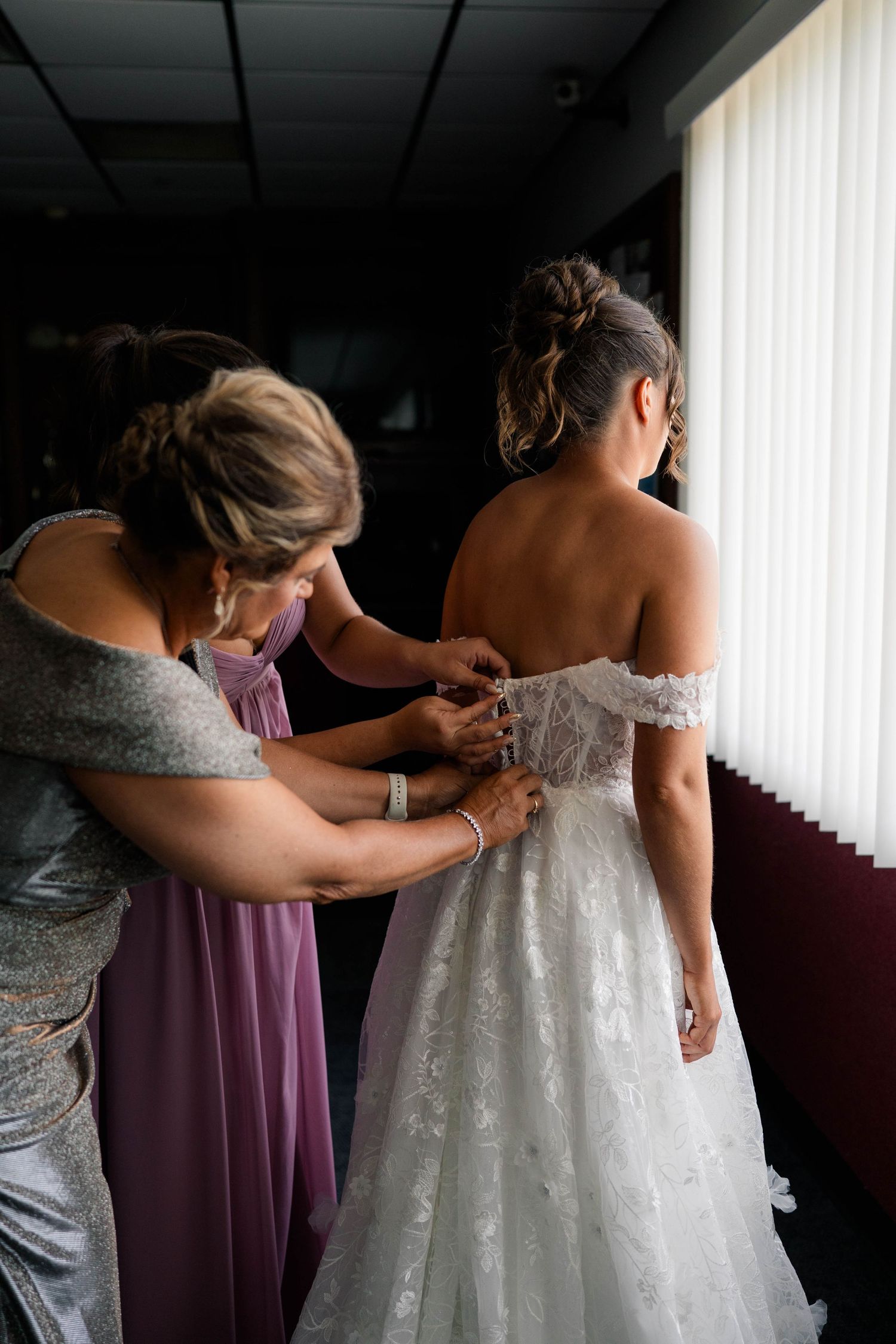 Two women helping a bride button her lace off-shoulder wedding dress from behind near a bright window.