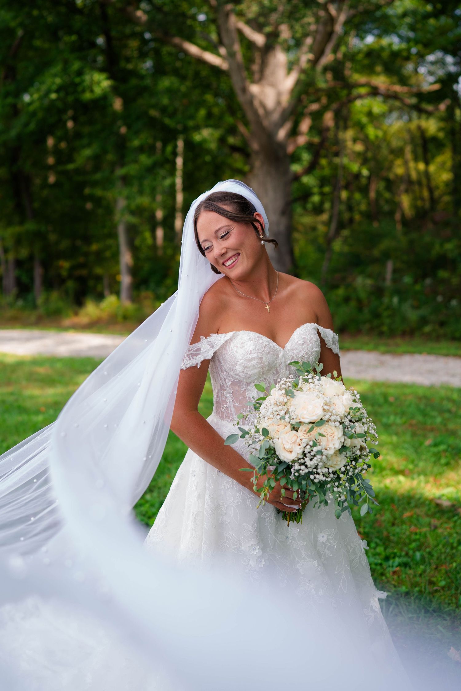 Smiling bride in lace off-shoulder gown with flowing veil holds white rose bouquet outdoors in green garden setting.