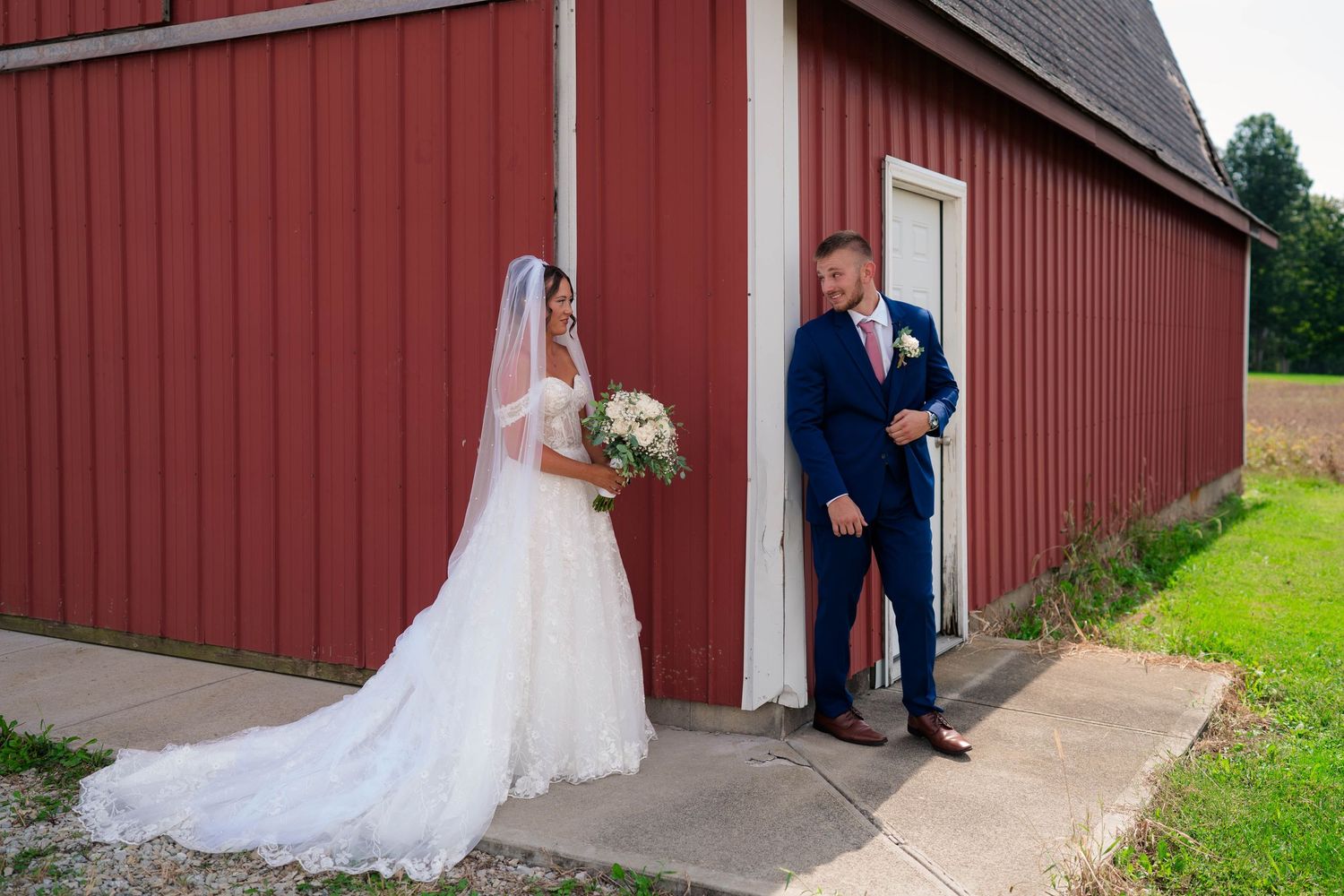 Groom seeing bride for first time in lace wedding gown during first look at red barn venue.