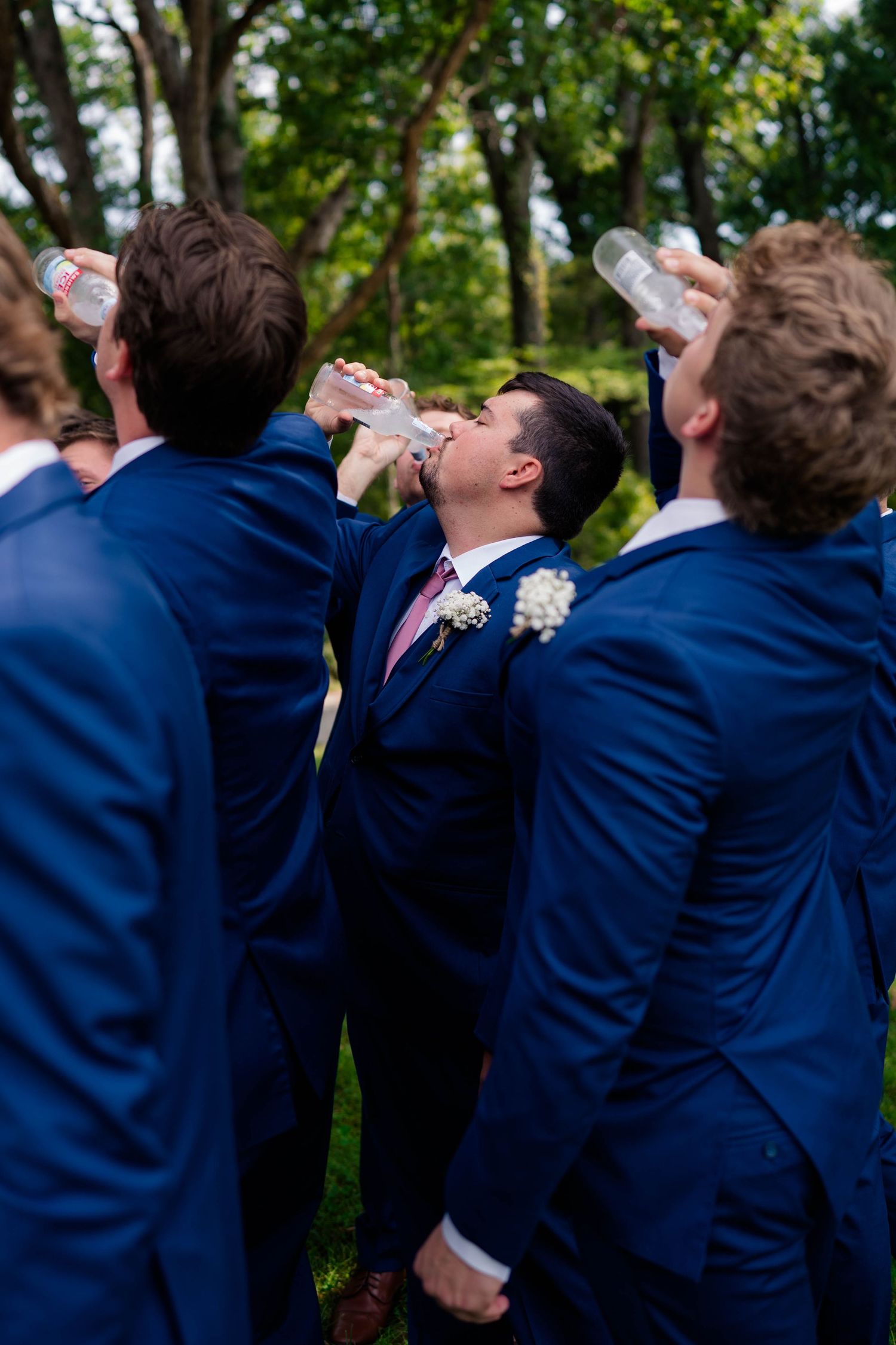 Groomsmen in navy suits humorously chug water bottles together outdoors at a wedding celebration.