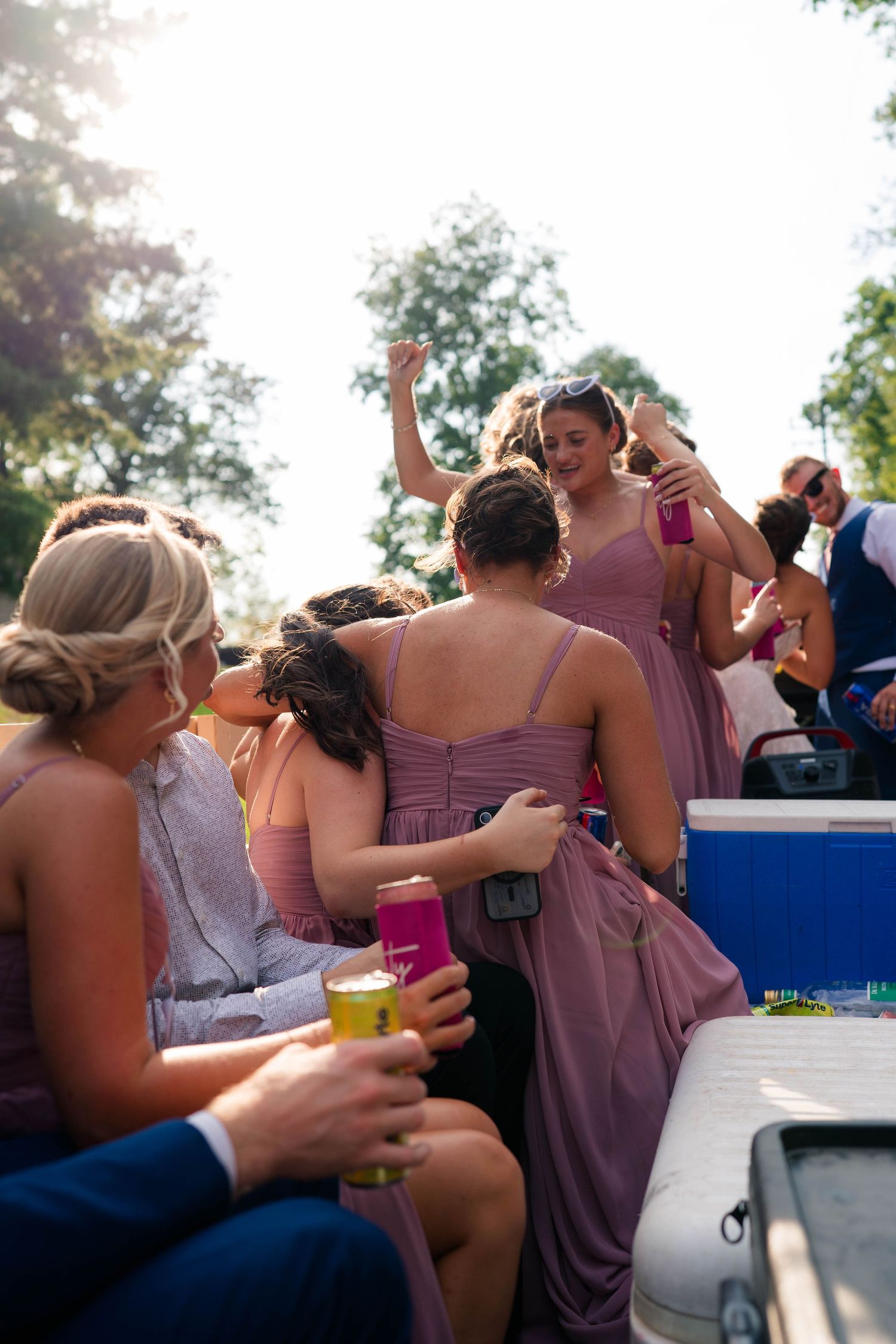 Bridesmaids in dusty rose dresses dancing and celebrating on truck bed at outdoor wedding.