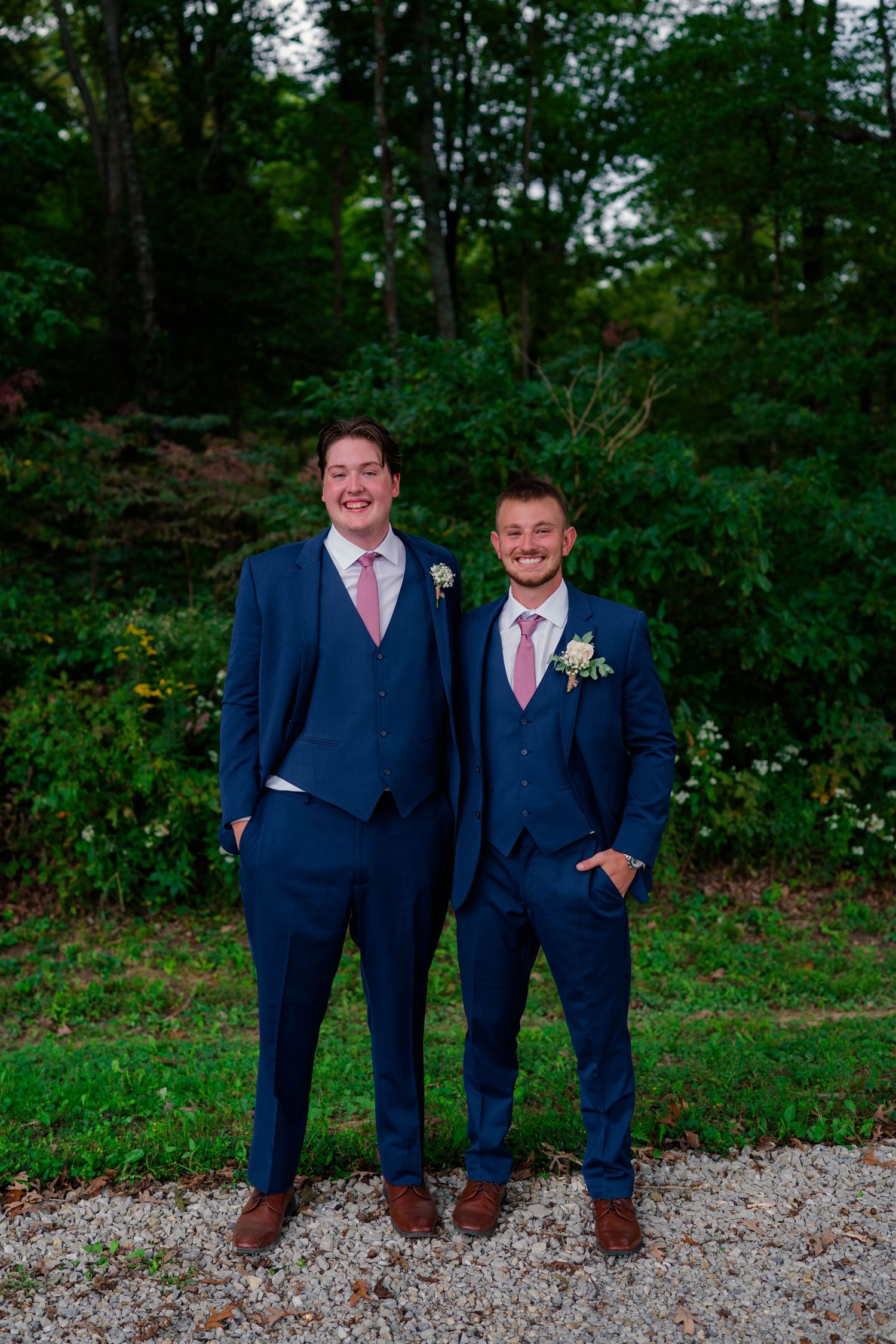 Two groomsmen smiling in navy blue suits with pink ties and boutonnieres at outdoor wedding.