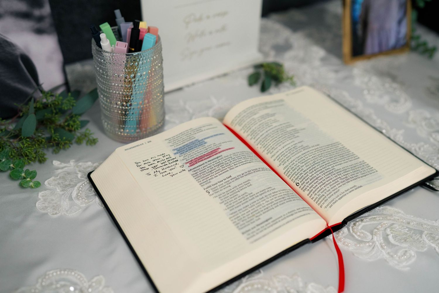 Open Bible with red ribbon bookmark displayed on lace-covered table with colorful pens at wedding reception.
