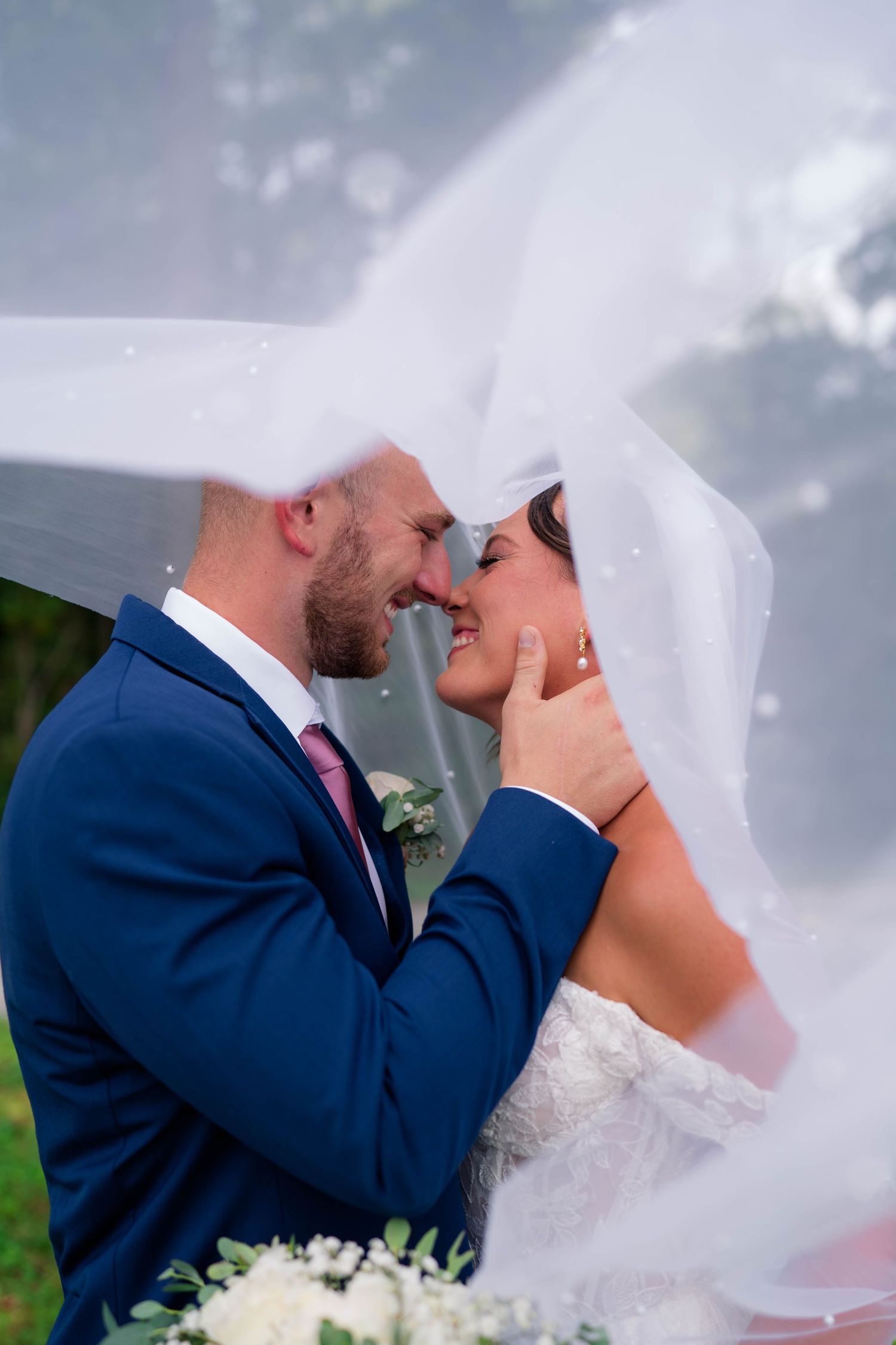 Romantic bride and groom nearly kissing beneath flowing white veil in an outdoor wedding portrait.