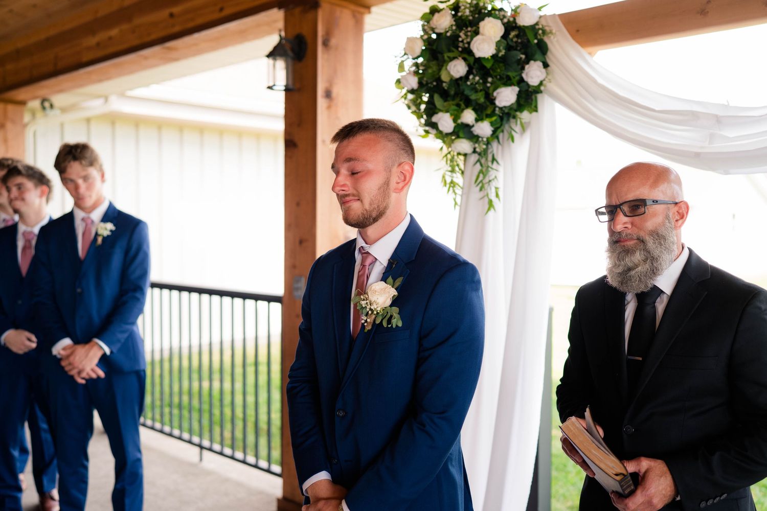 Groom in navy suit watches bride approach at floral arch altar with groomsmen and officiant.