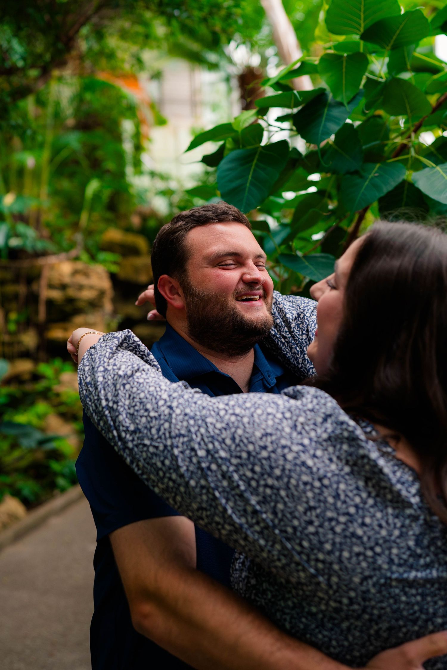 Joyful man smiling as his fiance embraces him in a lush tropical greenhouse setting.