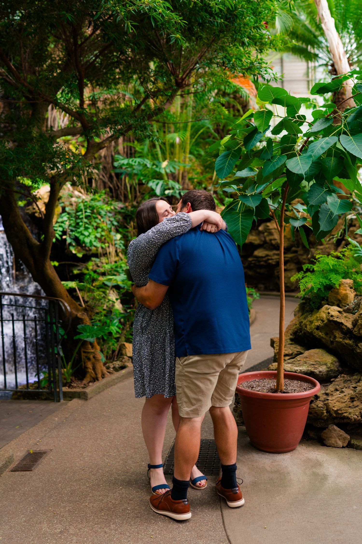 Couple embracing in a lush tropical greenhouse with waterfall and green plants.