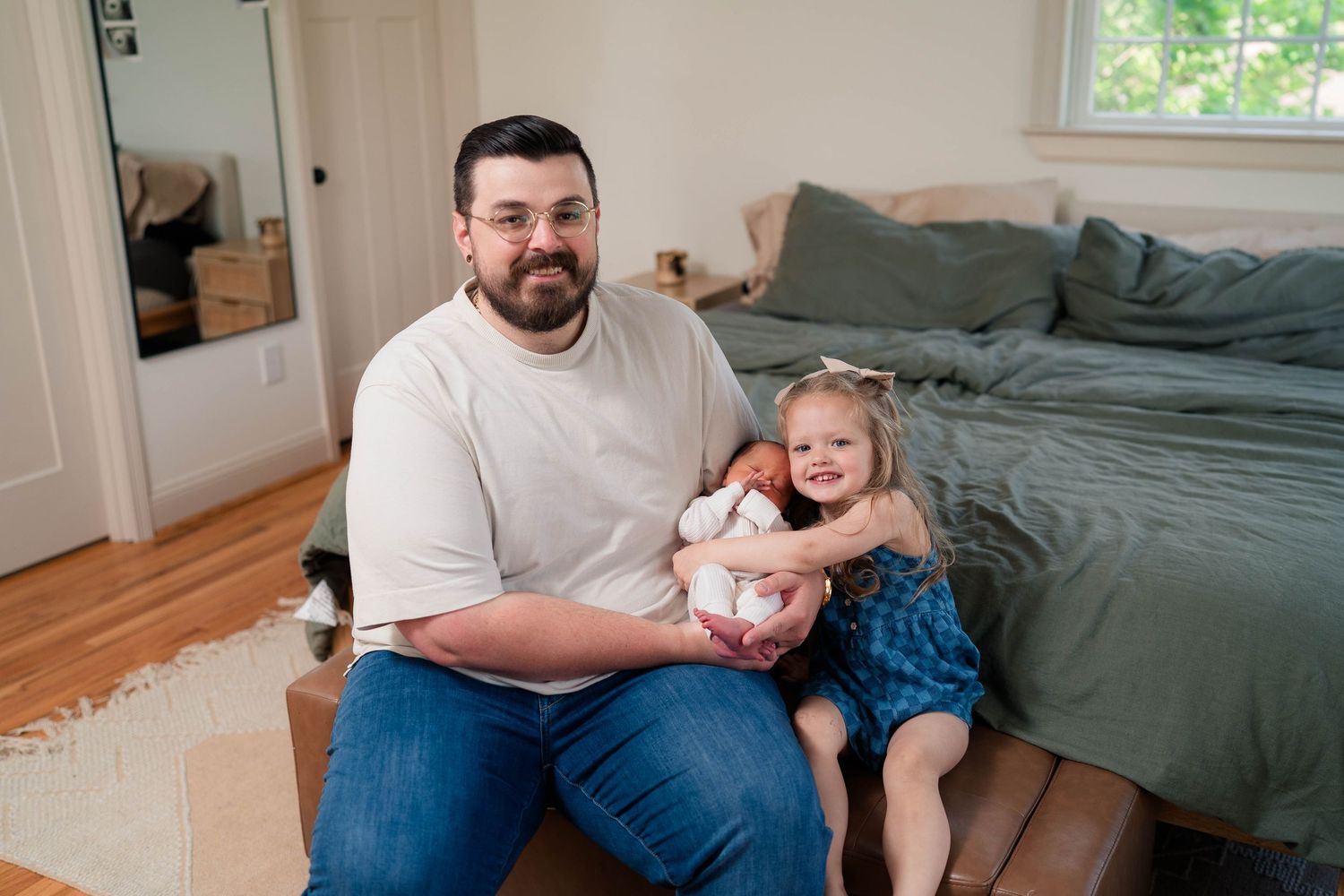 Smiling bearded father sits on ottoman with young daughter in blue bow hugging him in cozy bedroom.