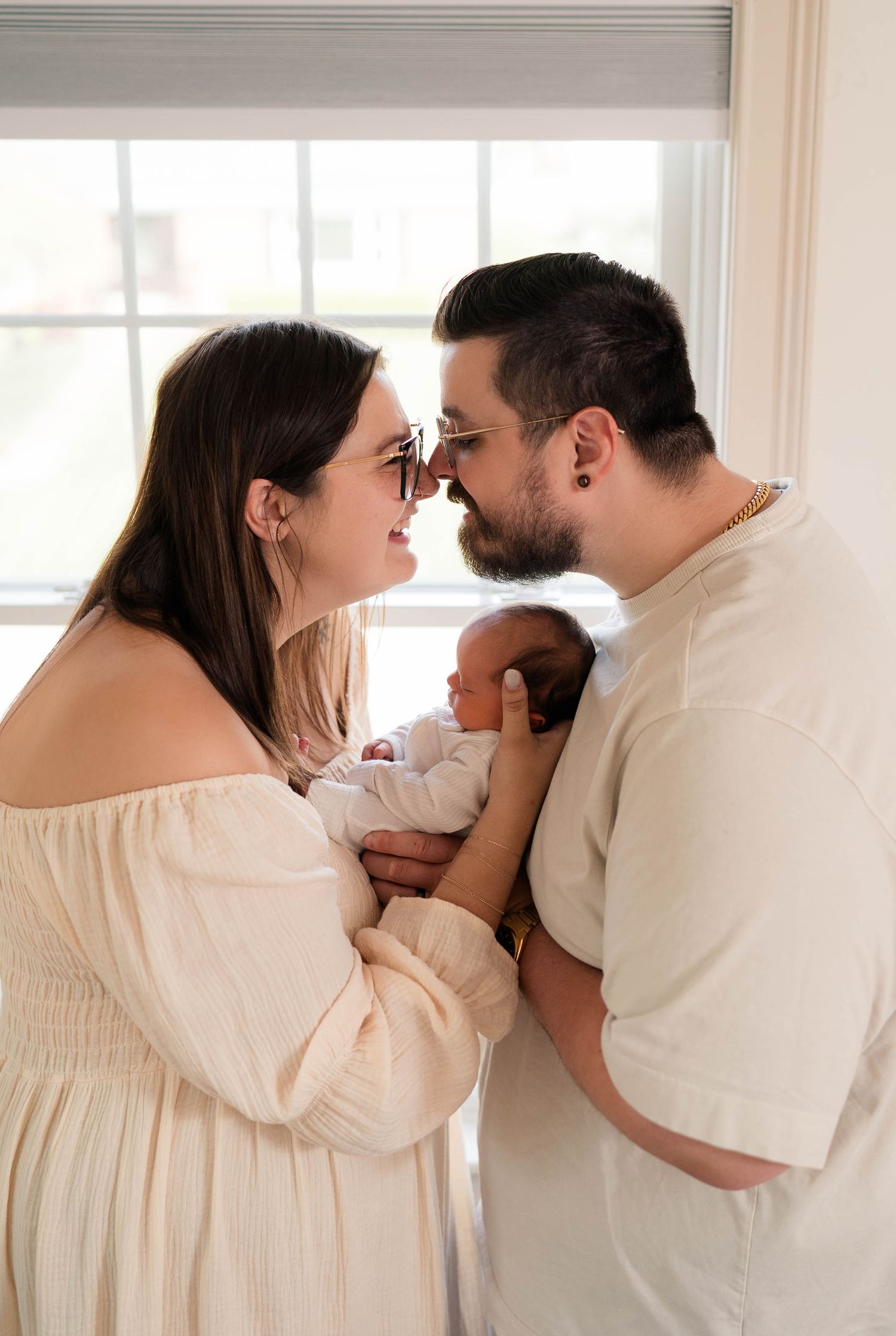 New parents touching foreheads while holding newborn baby together by window.