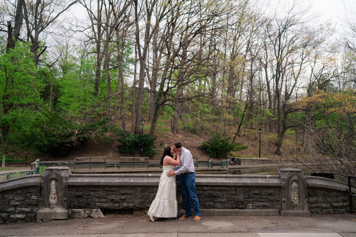 A couple shares a romantic kiss on a stone terrace surrounded by lush spring trees in a park.