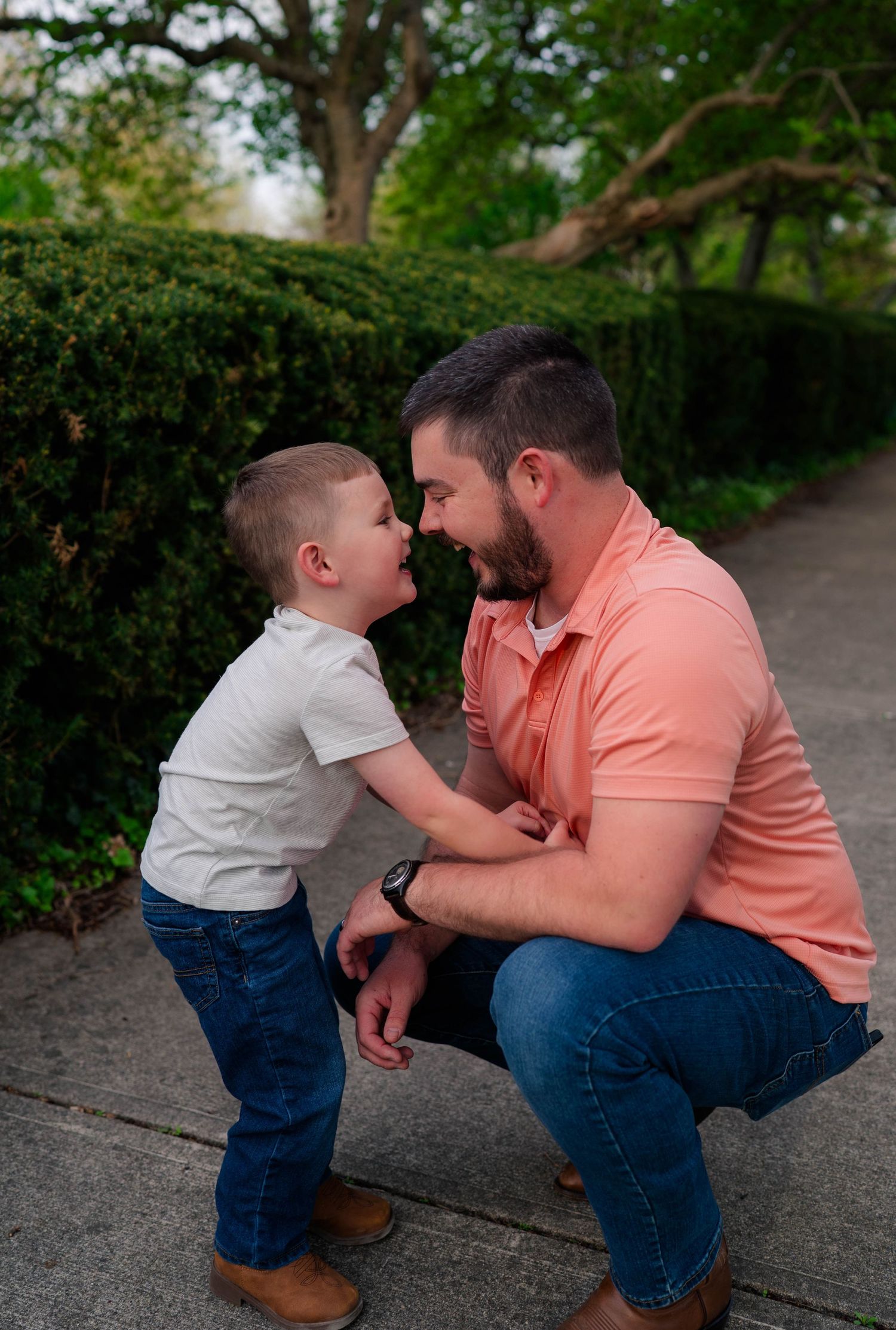 A father crouches down to share a sweet nose-to-nose moment with his young son outdoors in a lush green park.