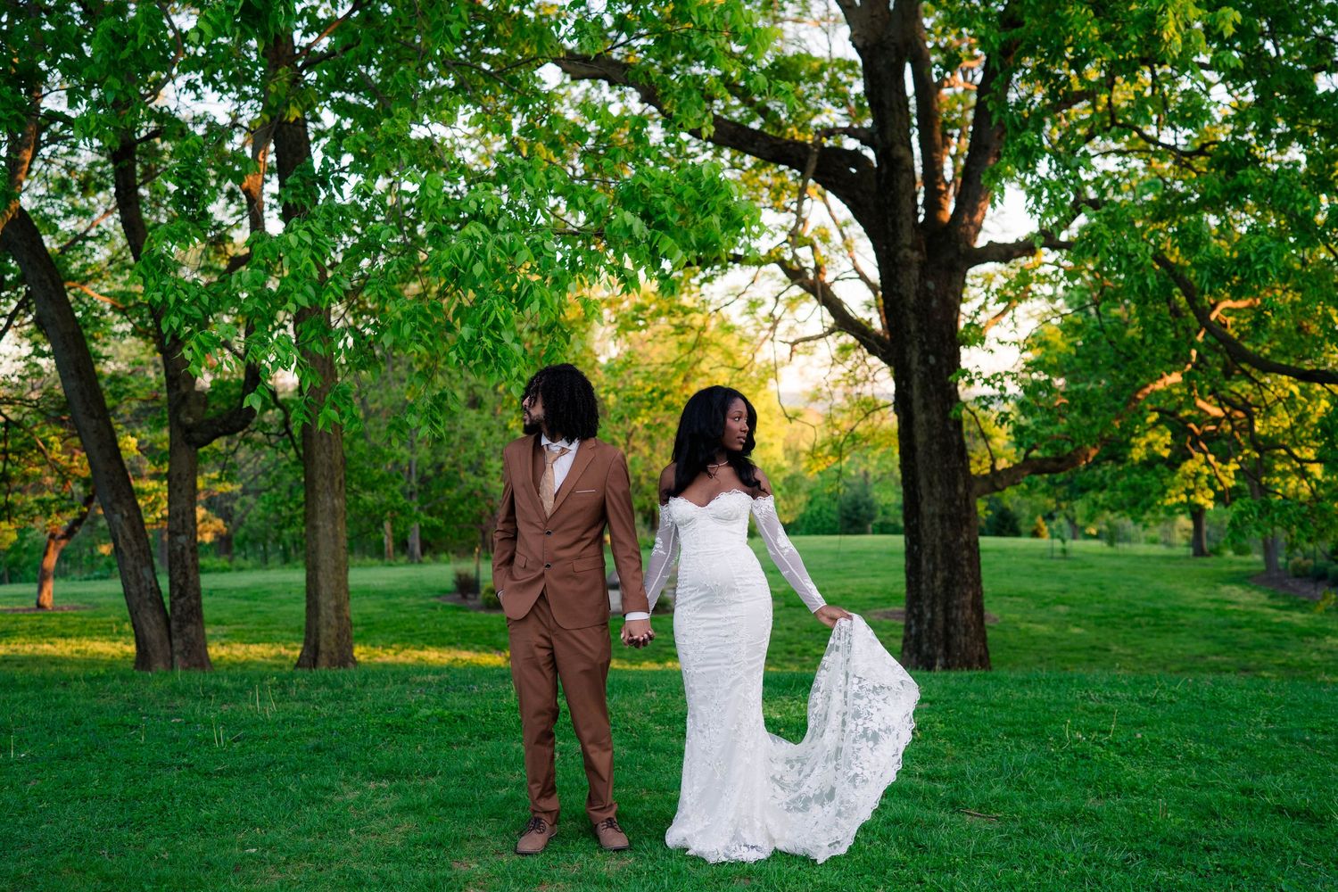 Bride in white lace gown and groom in tan suit hold hands in lush green park at golden hour.