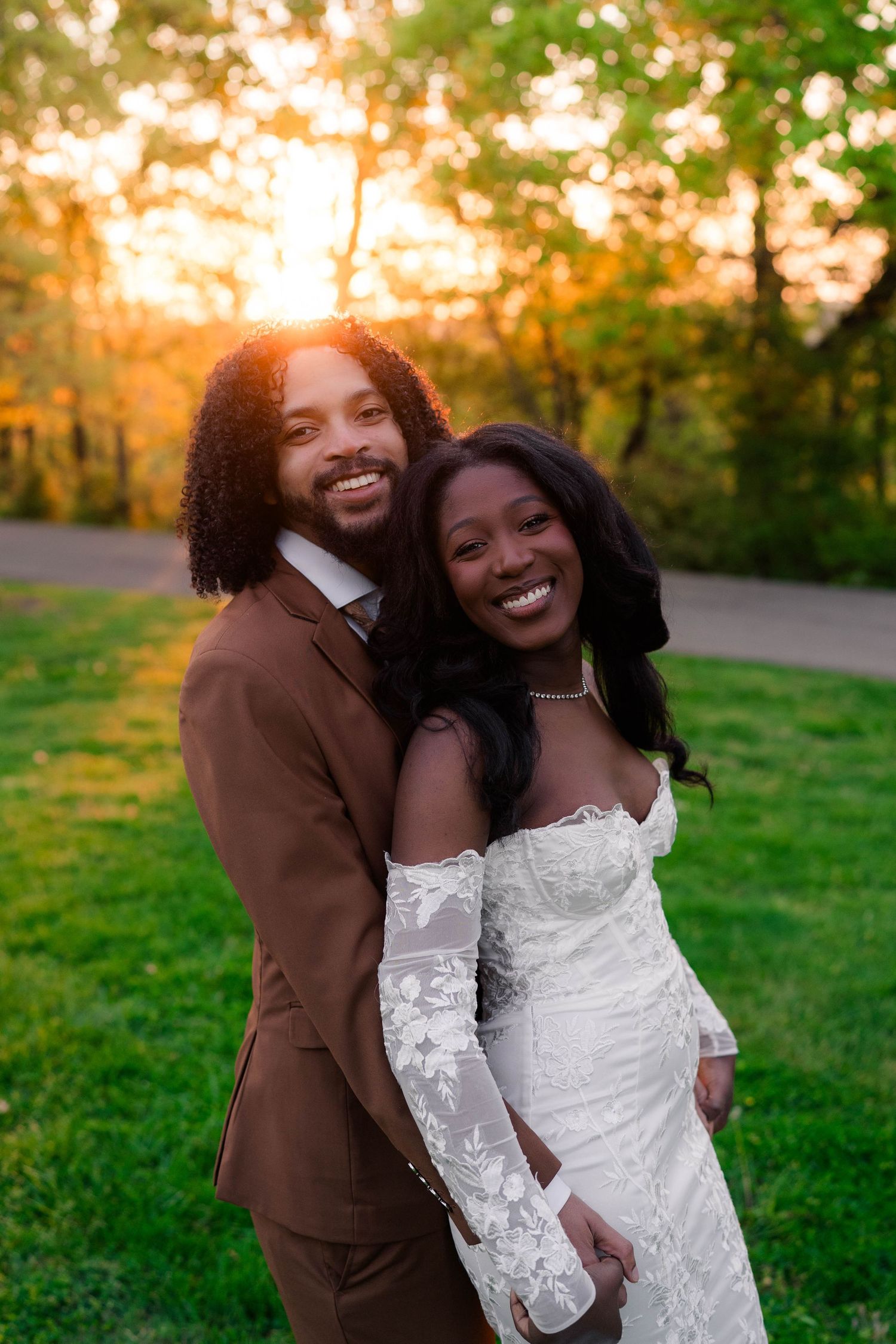 Smiling bride and groom pose outdoors at golden hour, she in lace gown, he in brown suit, surrounded by green trees.