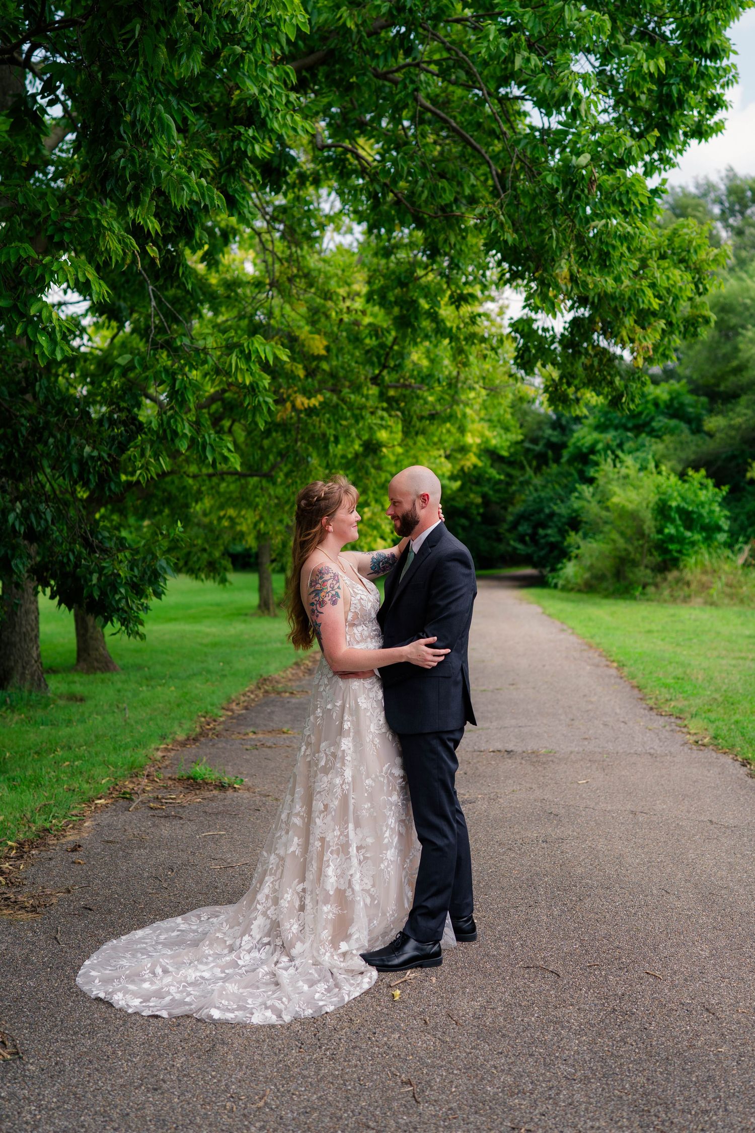 Bride and groom share a romantic embrace on a tree-lined path during their outdoor wedding portrait.