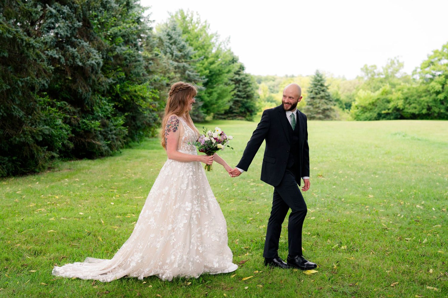 Groom in black tuxedo leads bride in lace ballgown across green lawn at outdoor wedding.