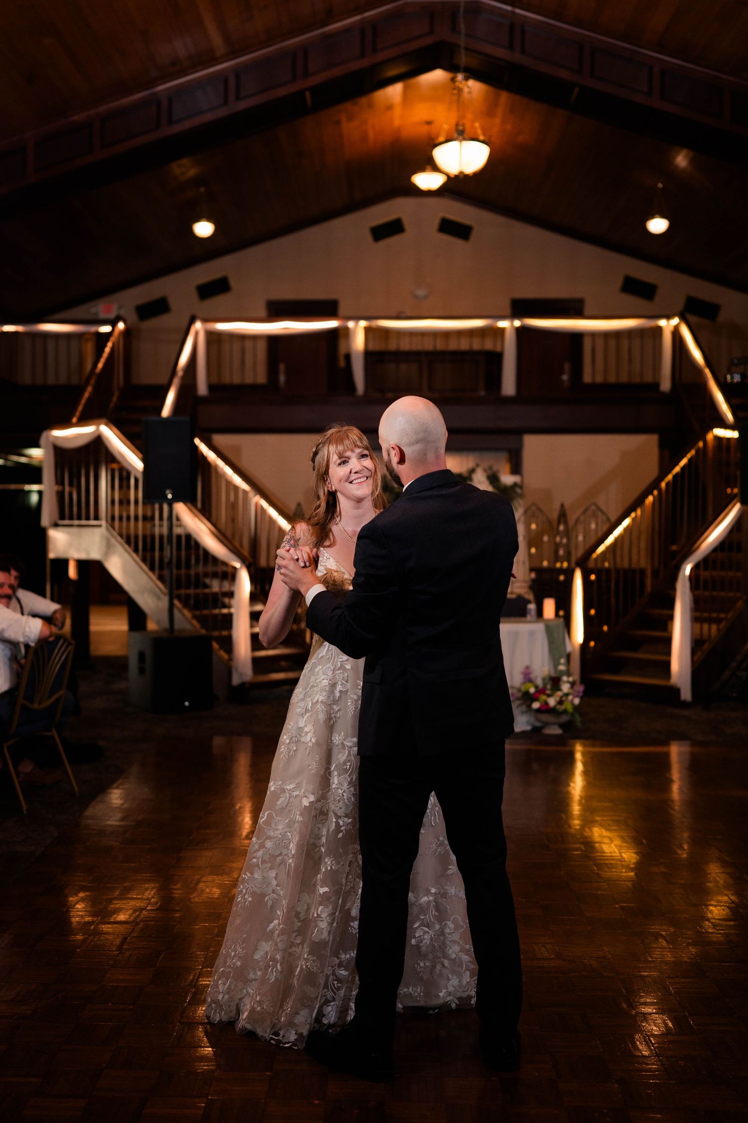 Bride and groom share first dance on wooden dance floor in warmly lit wedding reception venue.