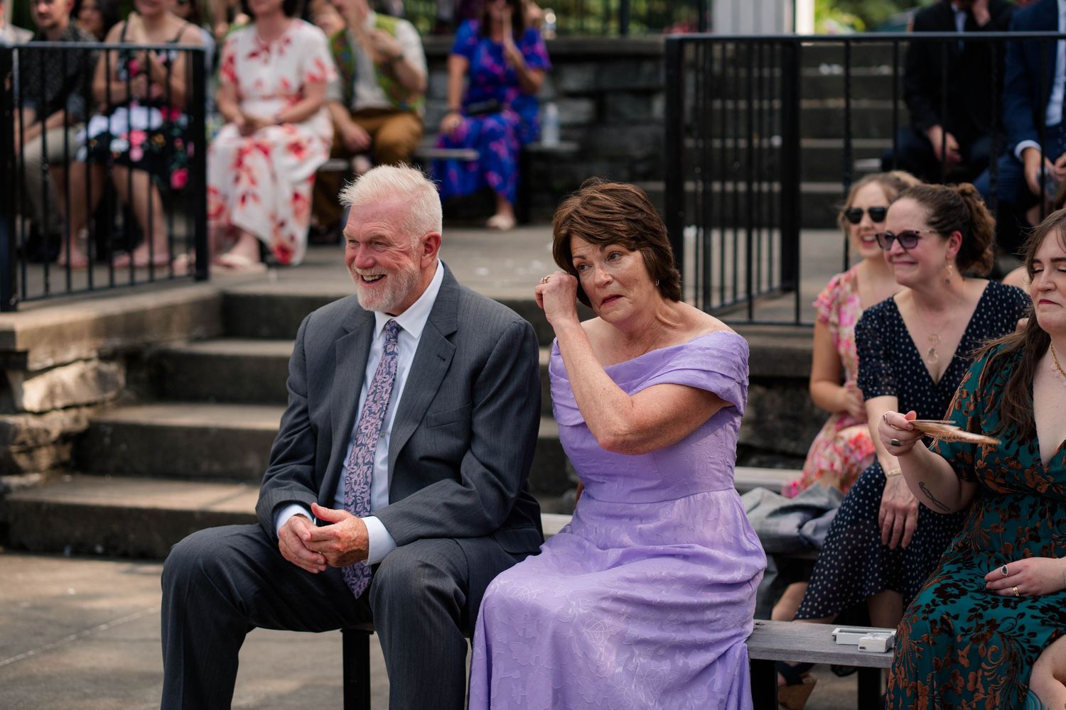 Couple in formal attire seated as emotional guests at outdoor wedding.