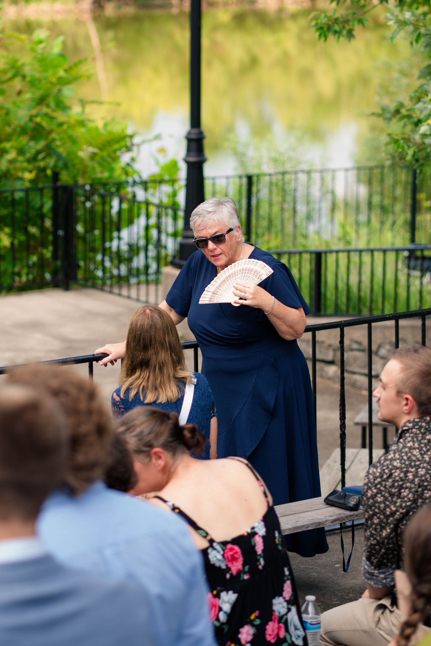 Elderly woman in navy dress holding decorative fan outdoors near pond at wedding ceremony.